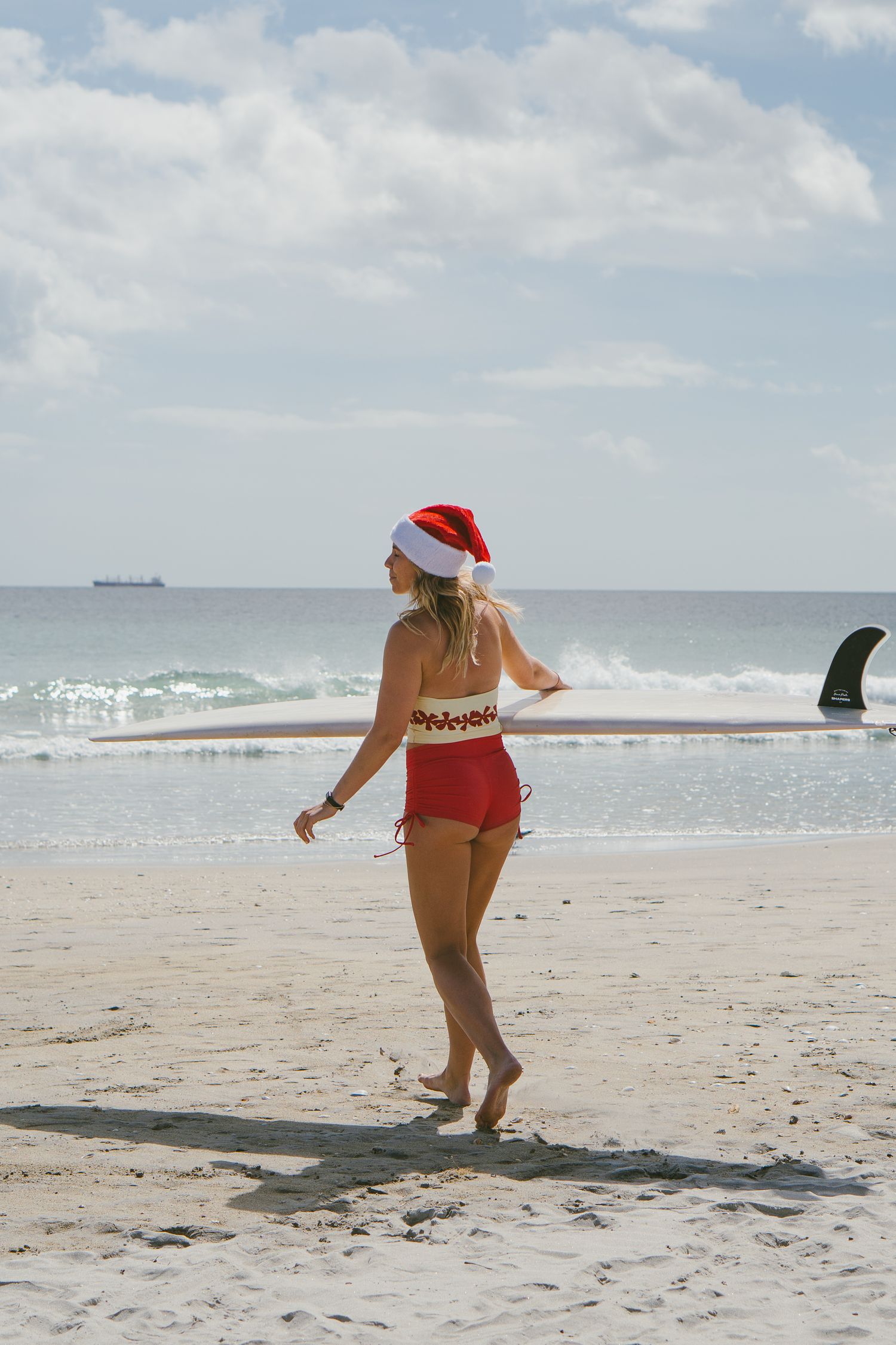 Person in Santa hat walking along a sunny beach shoreline.