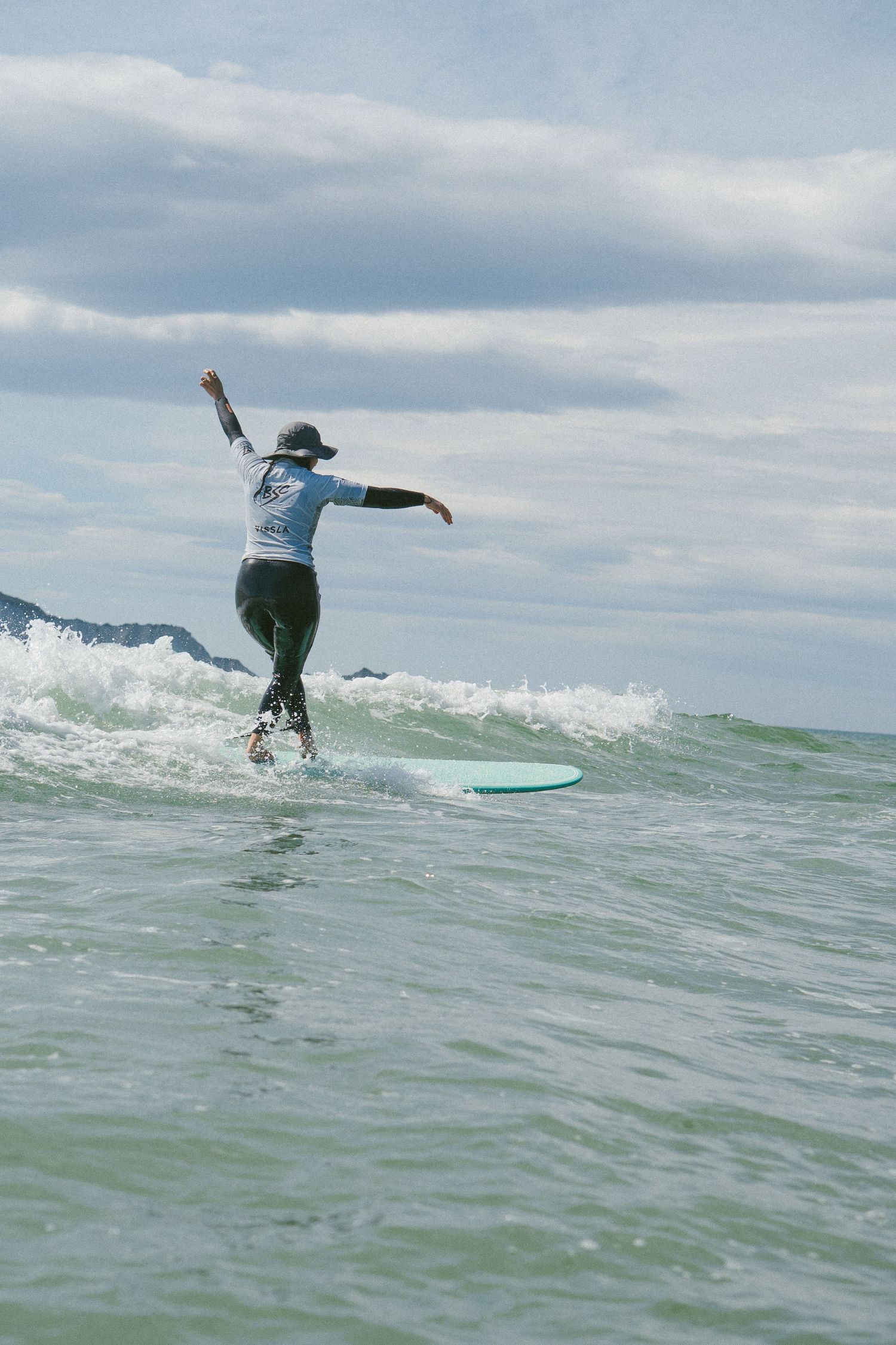 Surfer rides a turquoise wave while wearing a blue shirt and hat on a cloudy day at sea.