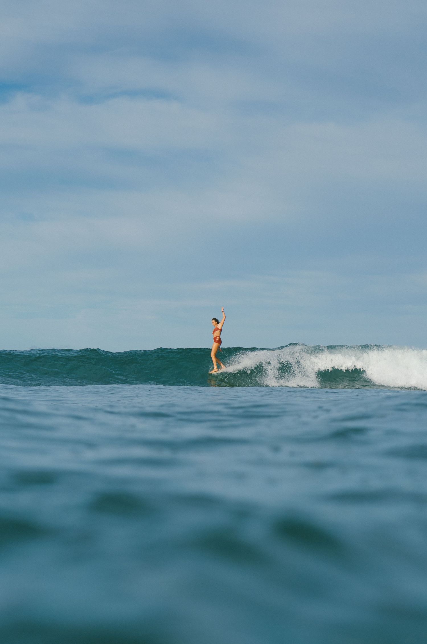 A surfer in a yellow bikini rides a wave on a longboard in the open ocean under a cloudy blue sky.