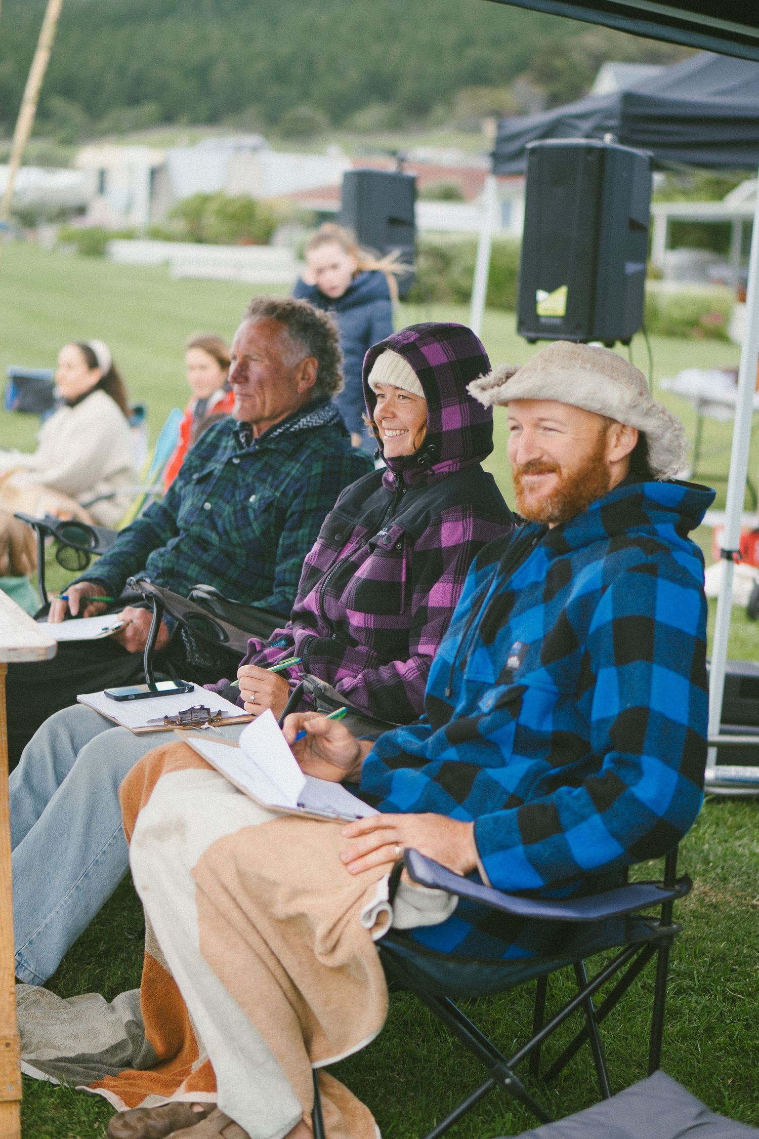Spectators sitting in camping chairs under a tent watching an outdoor event.