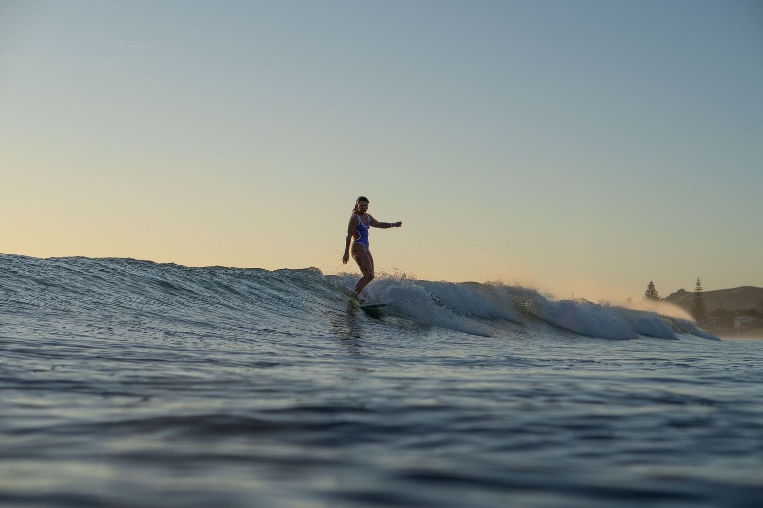 Surfer rides along the face of a wave during sunset with ocean spray trailing behind.