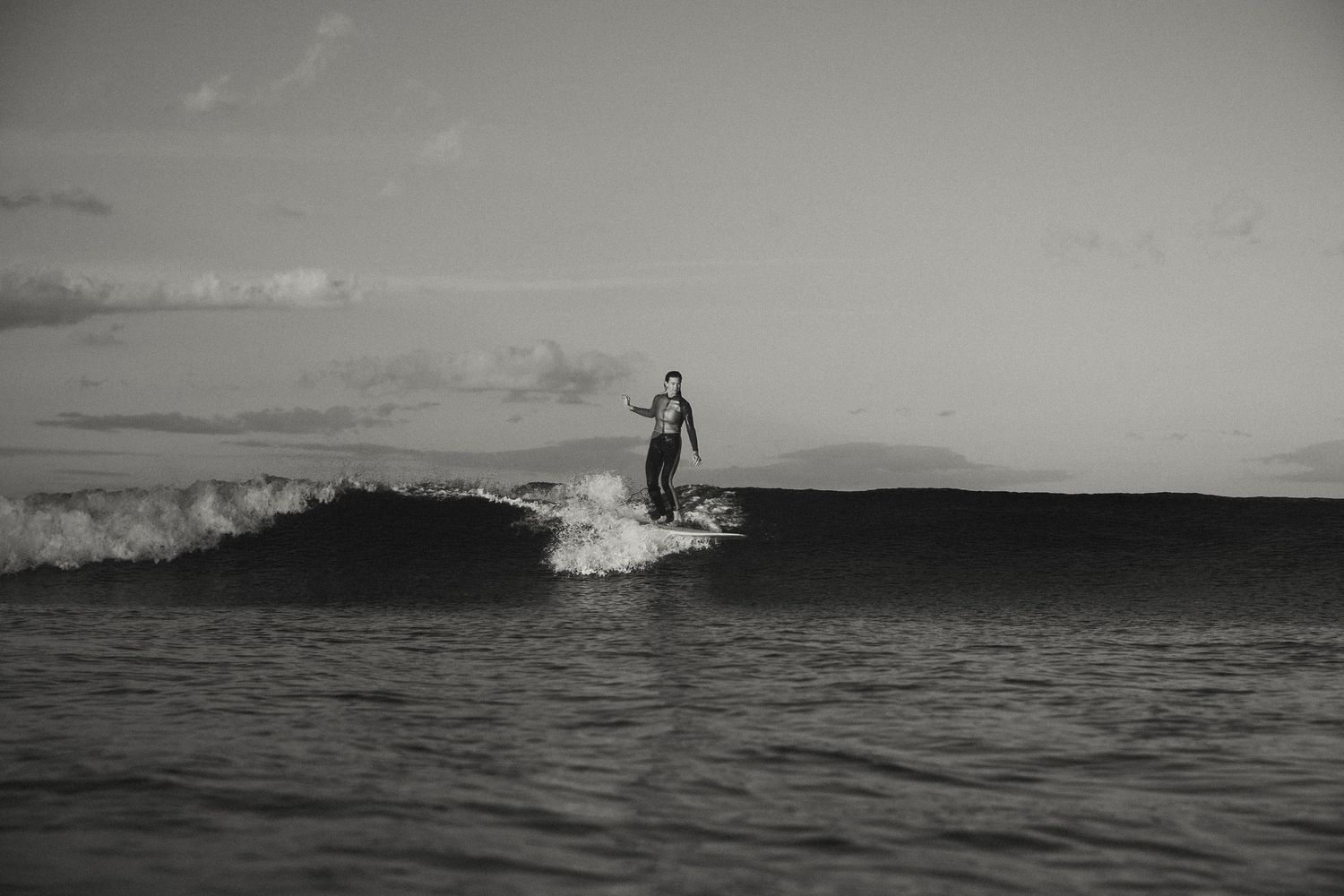Surfer silhouette standing on board against dramatic ocean waves at sunset.