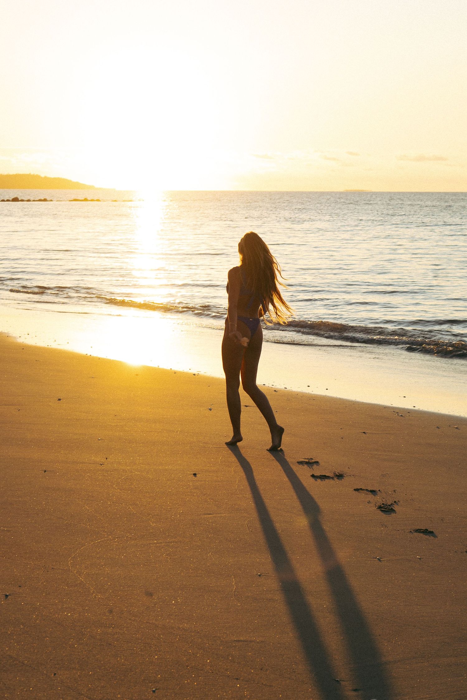 Silhouette walks along empty beach at golden sunset casting long shadow on sand.