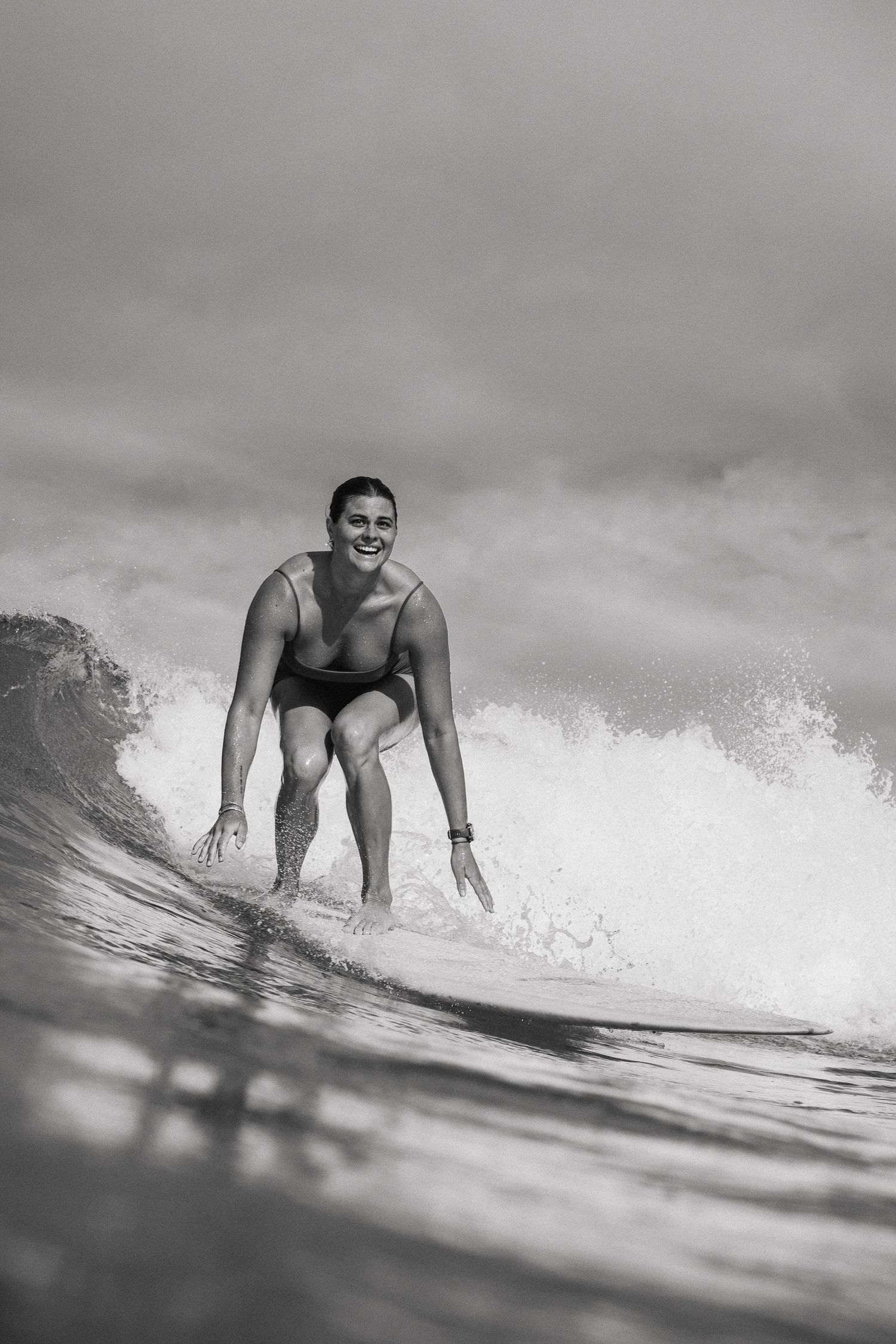 Black and white photo of a smiling surfer riding a wave in the ocean on a cloudy day.