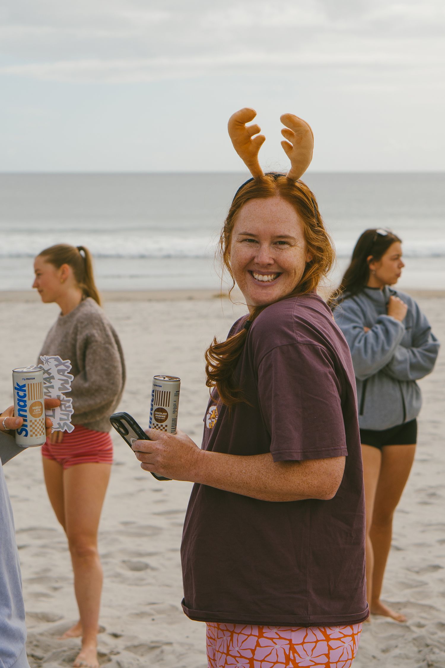Friends enjoying drinks and wearing reindeer antler headbands on a cloudy beach day.