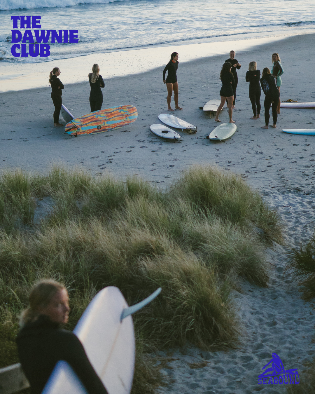 Group of surfers with boards on a beach at dusk, gathered near dunes for The Dawnie Club surf session.