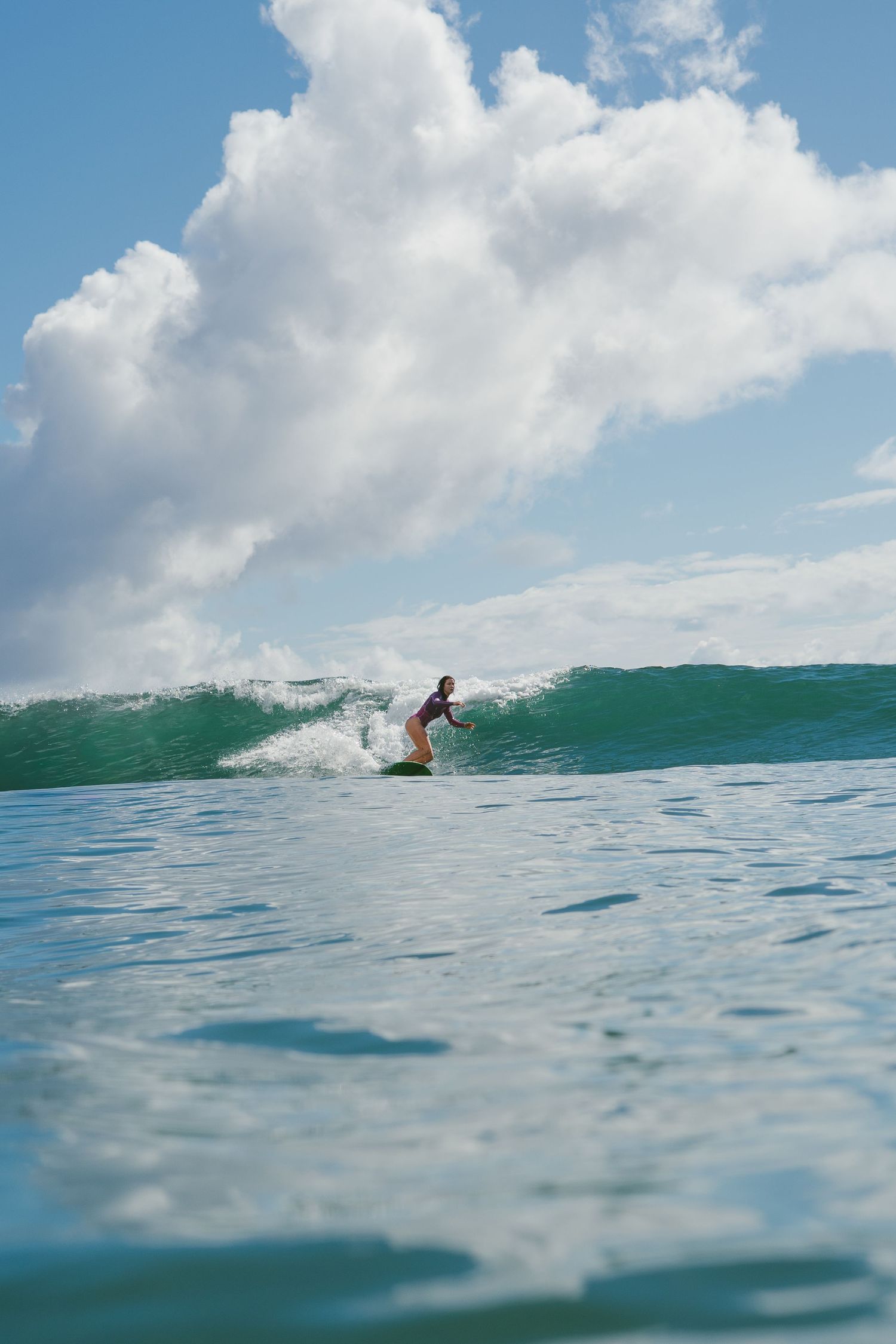 Distant surfer catches a perfect emerald wave under fluffy white clouds.