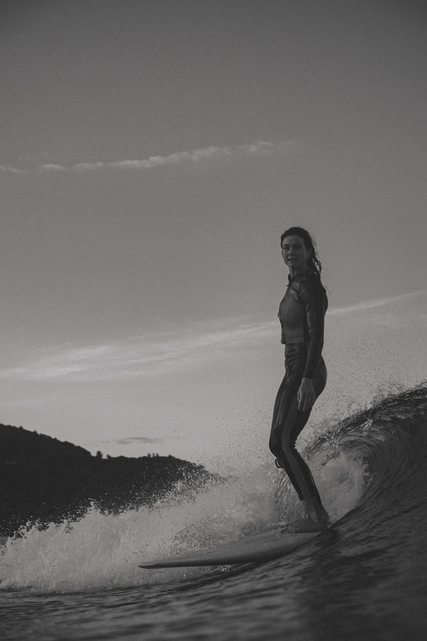 Surfer riding a breaking wave in black and white against coastal backdrop.