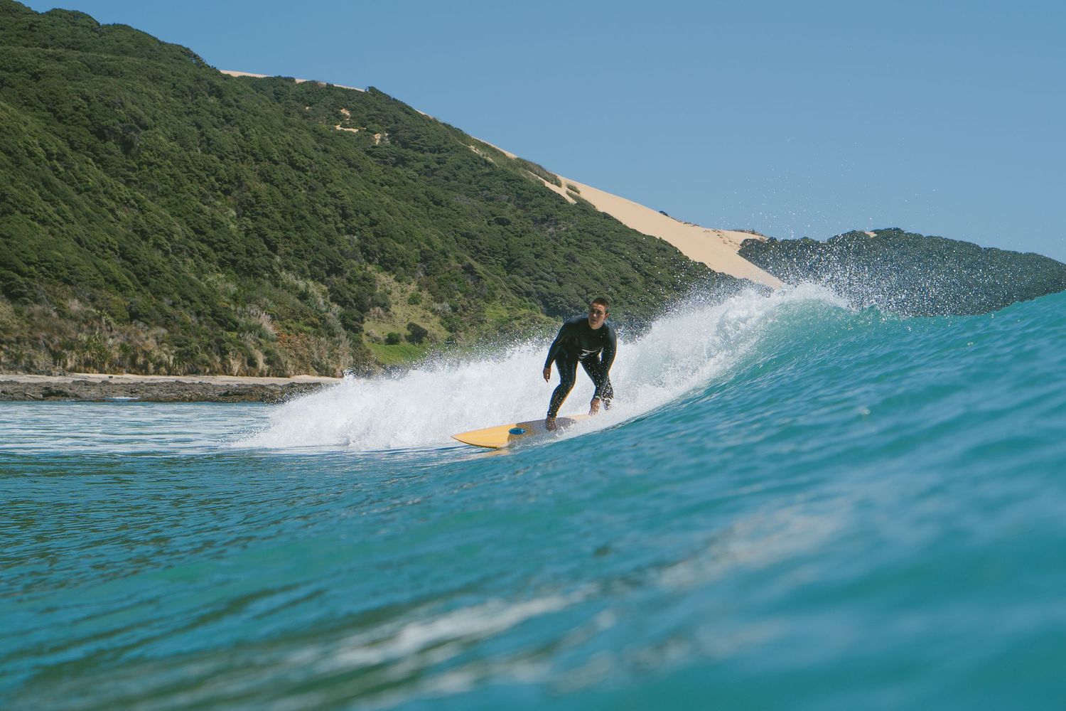 Surfer rides a breaking wave in crystal clear turquoise waters along a scenic coastline.