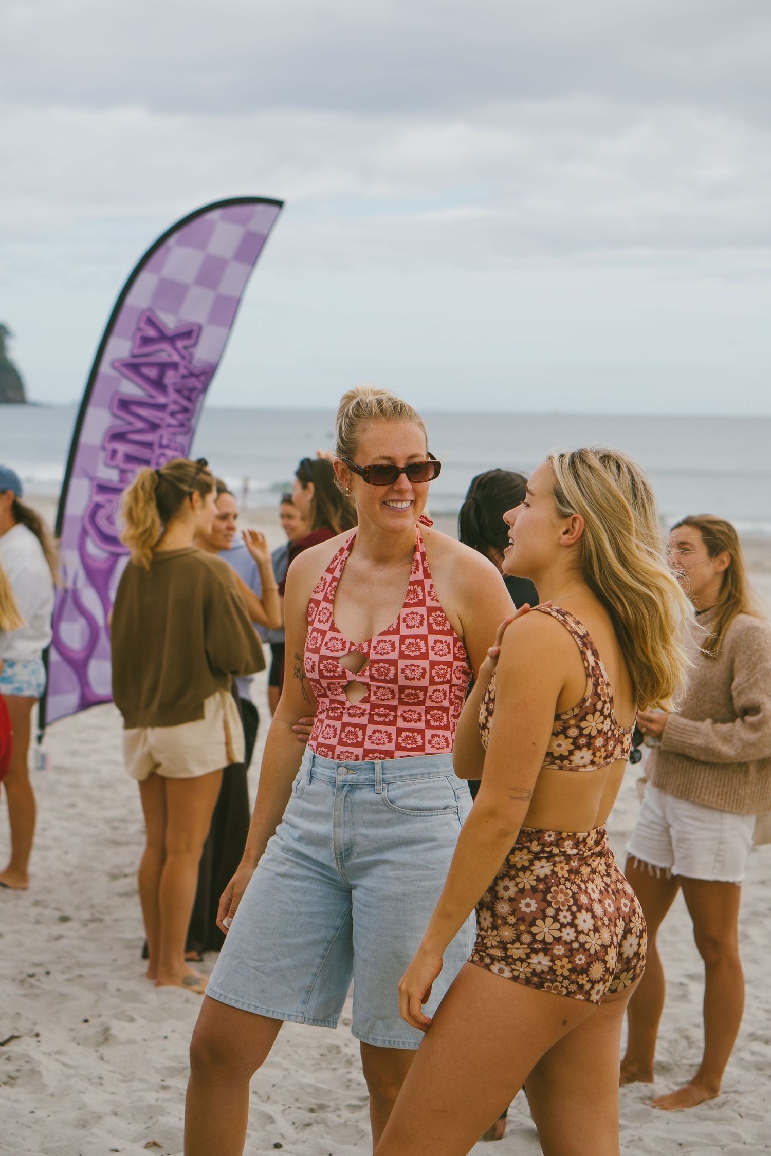 Group in retro-style swimwear gather on beach near purple banner flags.
