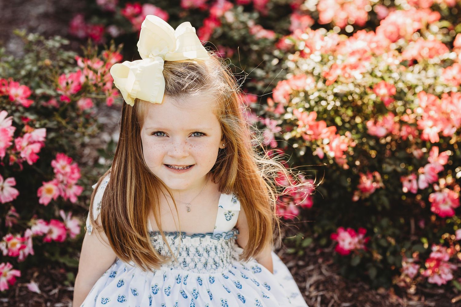 A young child in a blue dress and white bow poses in front of blooming pink flowers.