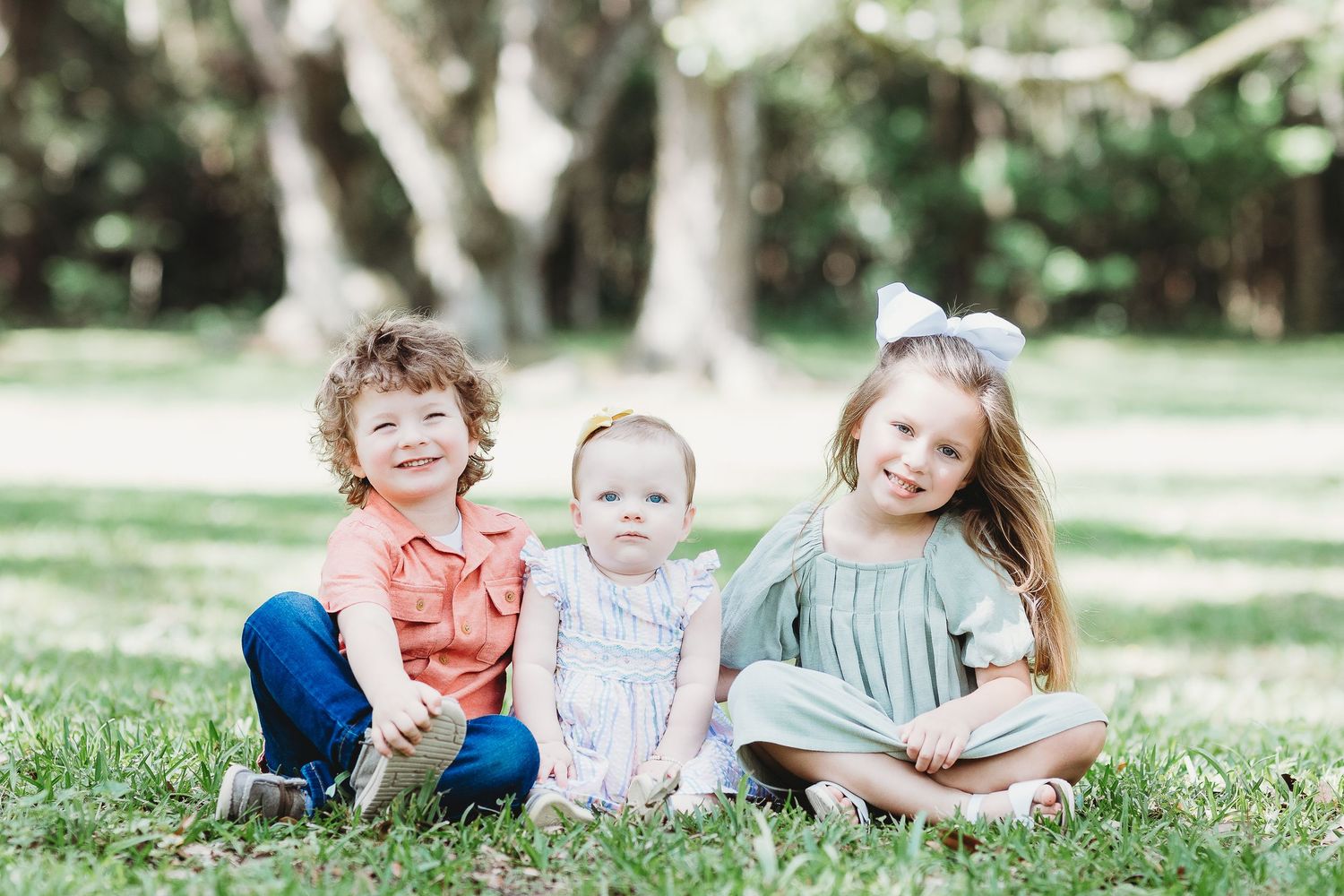 Three young children sit together on grass under shady trees.