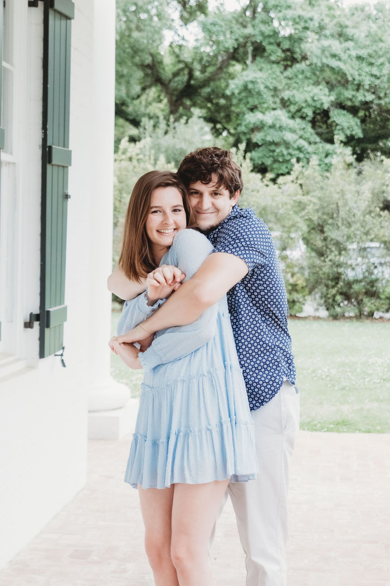 A couple embraces on a white porch with green foliage in the background.