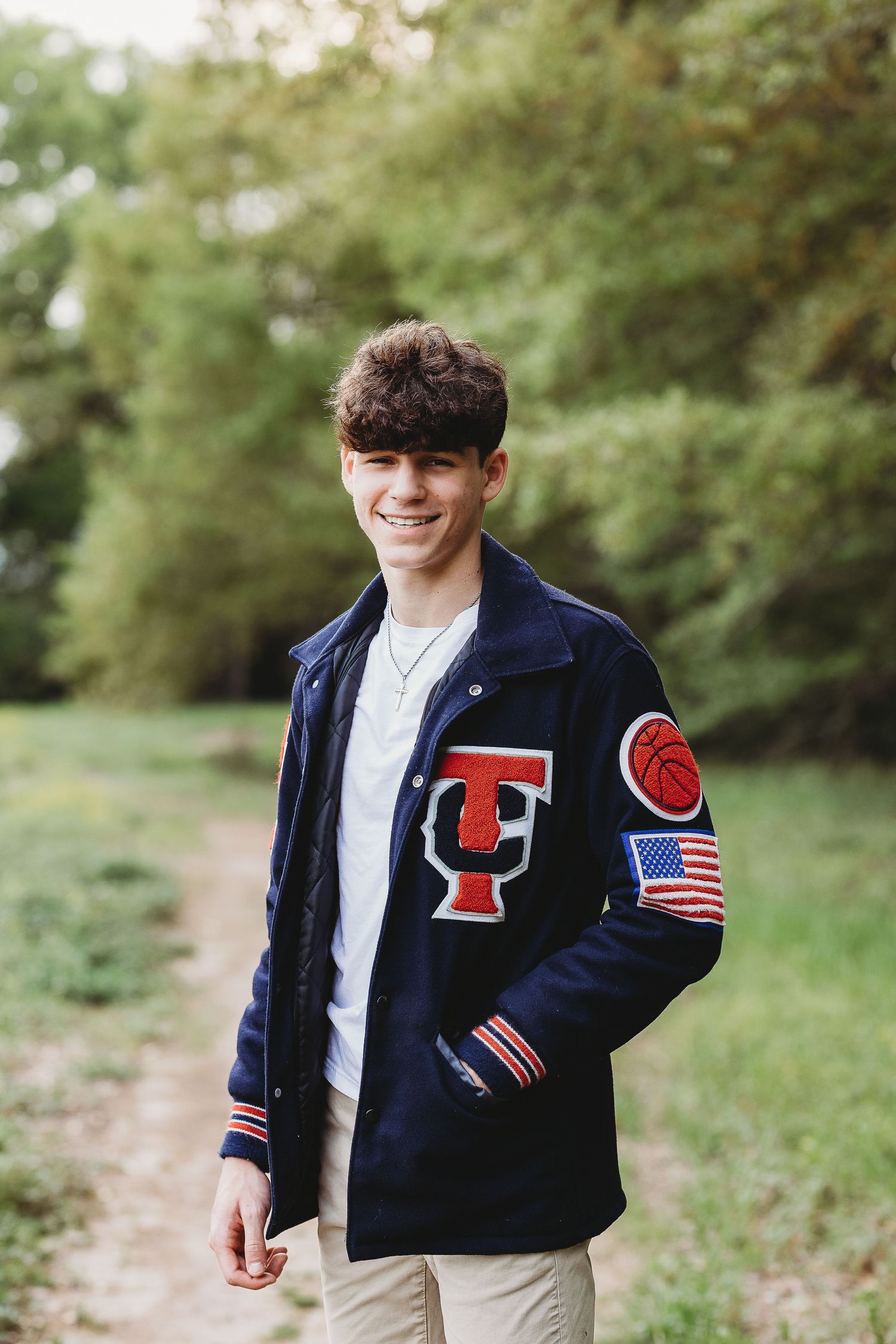 Student wearing a navy varsity letterman jacket with patches and American flag emblem poses outdoors in natural setting.