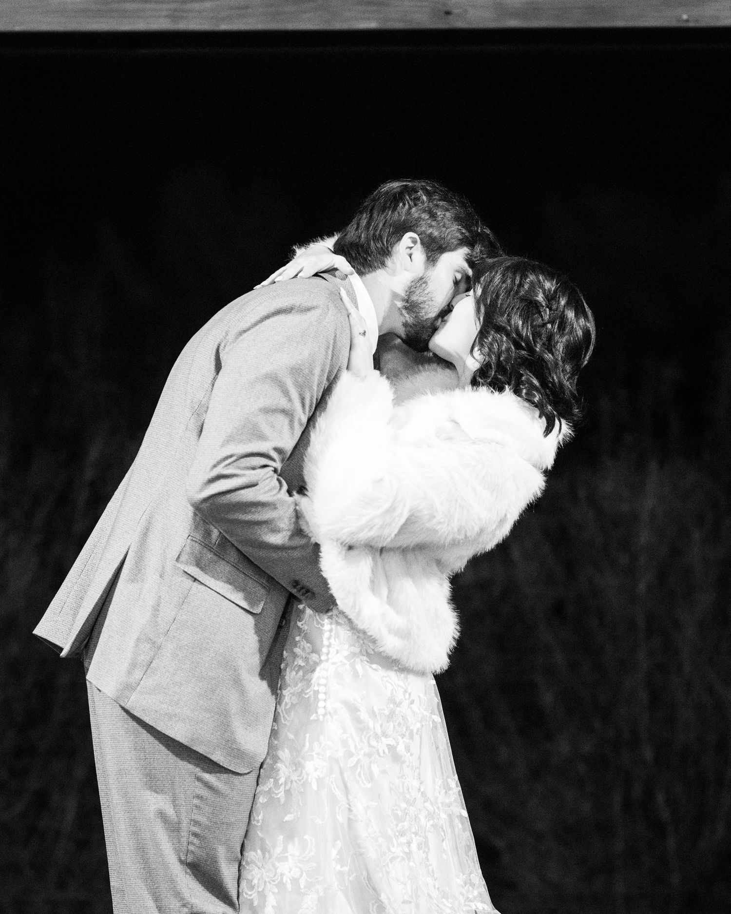 Black and white wedding photo of a romantic kiss between a couple, with the bride wearing a fur shawl and lace dress.