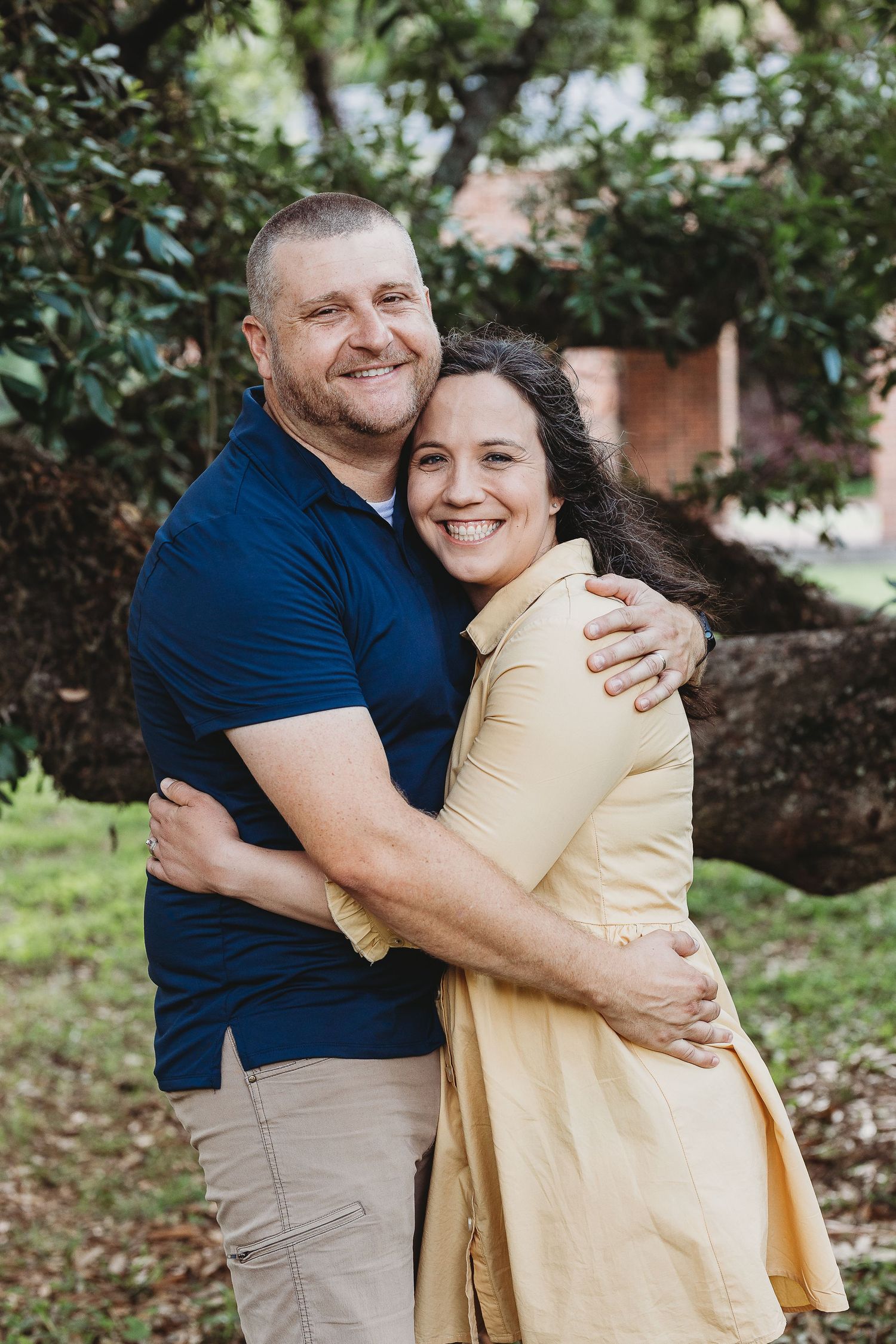 Couple embraces while smiling in casual attire under green trees.