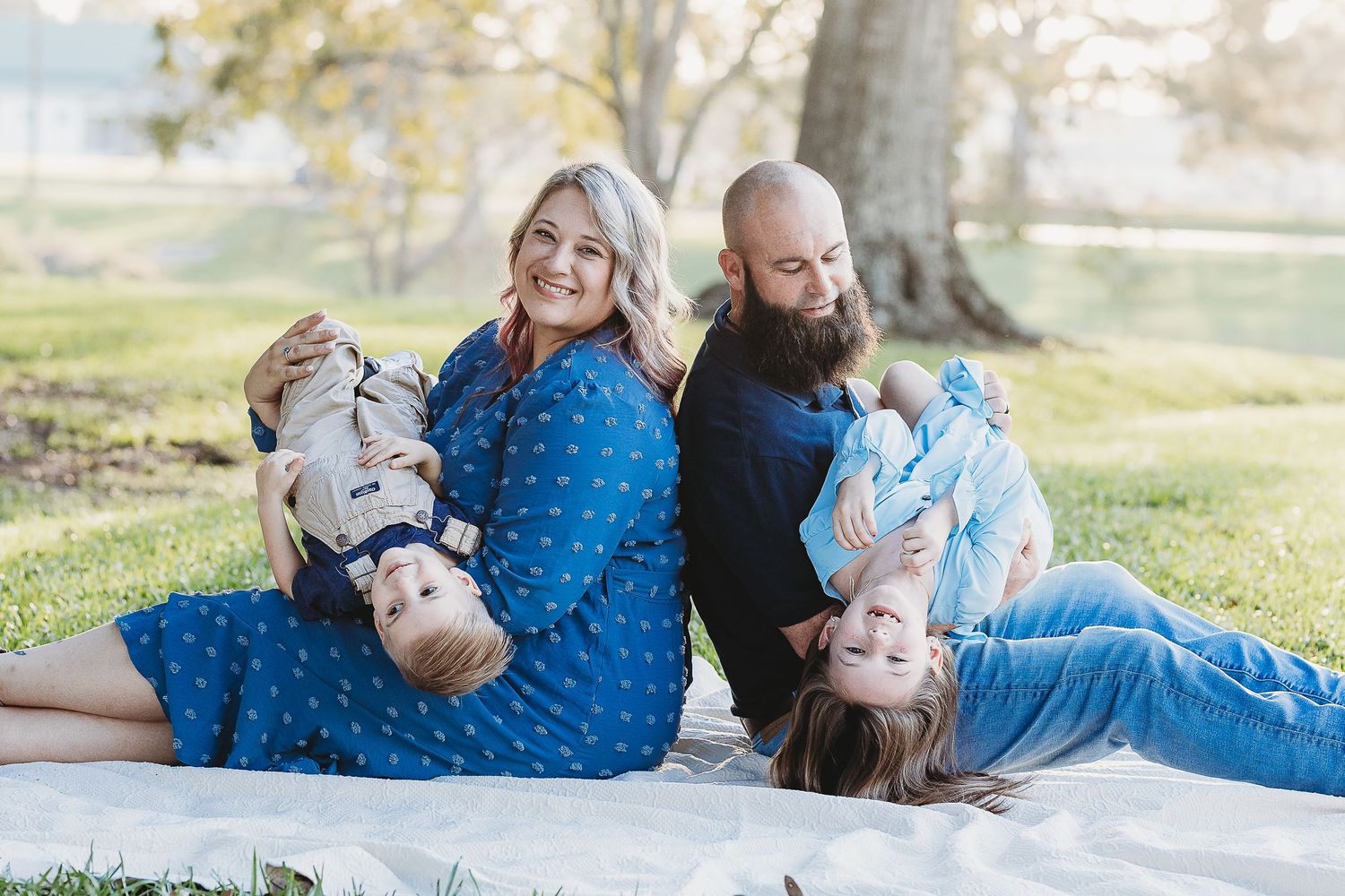 A family plays and laughs together while lying on a white blanket in a grassy park.