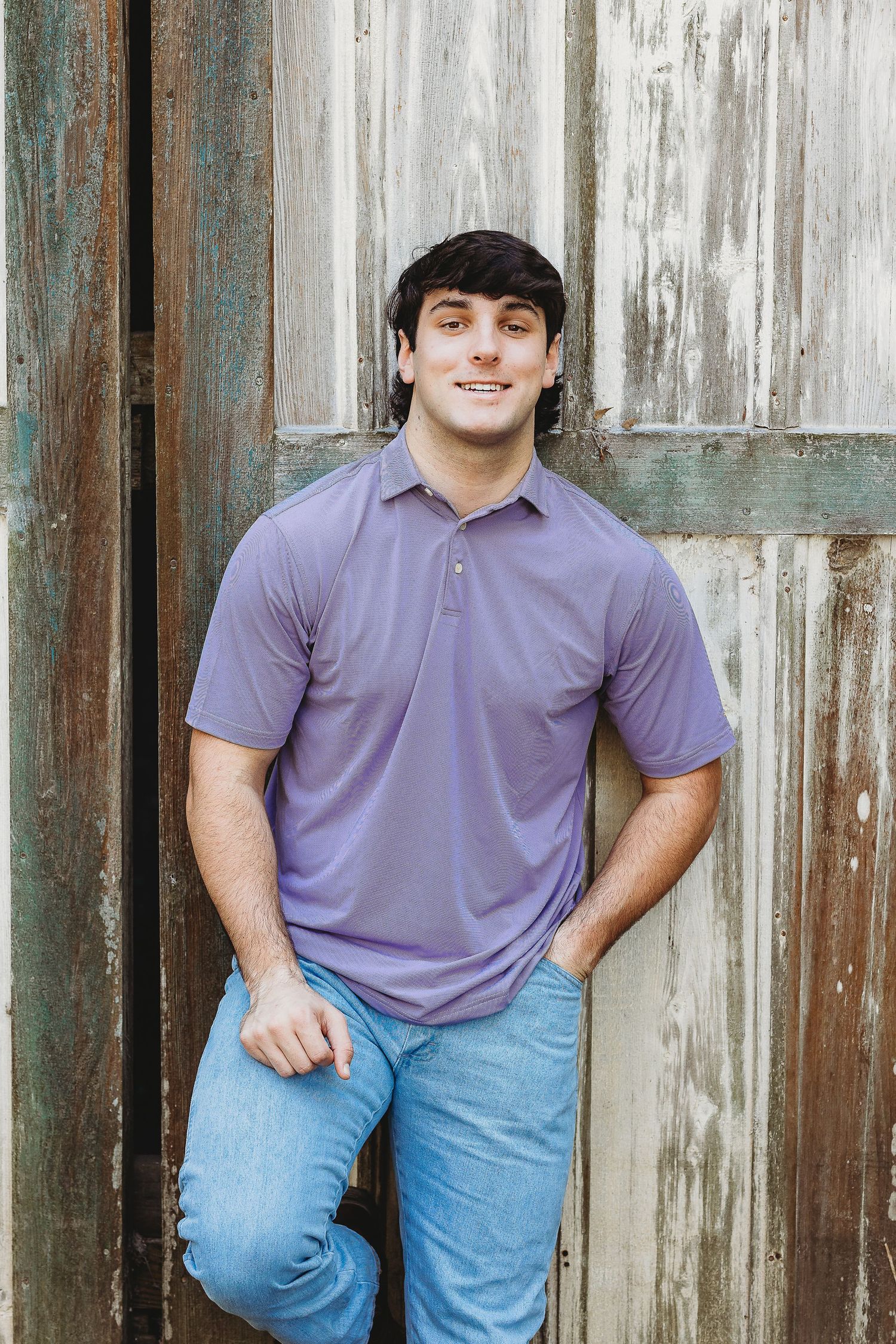 Portrait against weathered wooden wall wearing purple polo shirt and jeans.