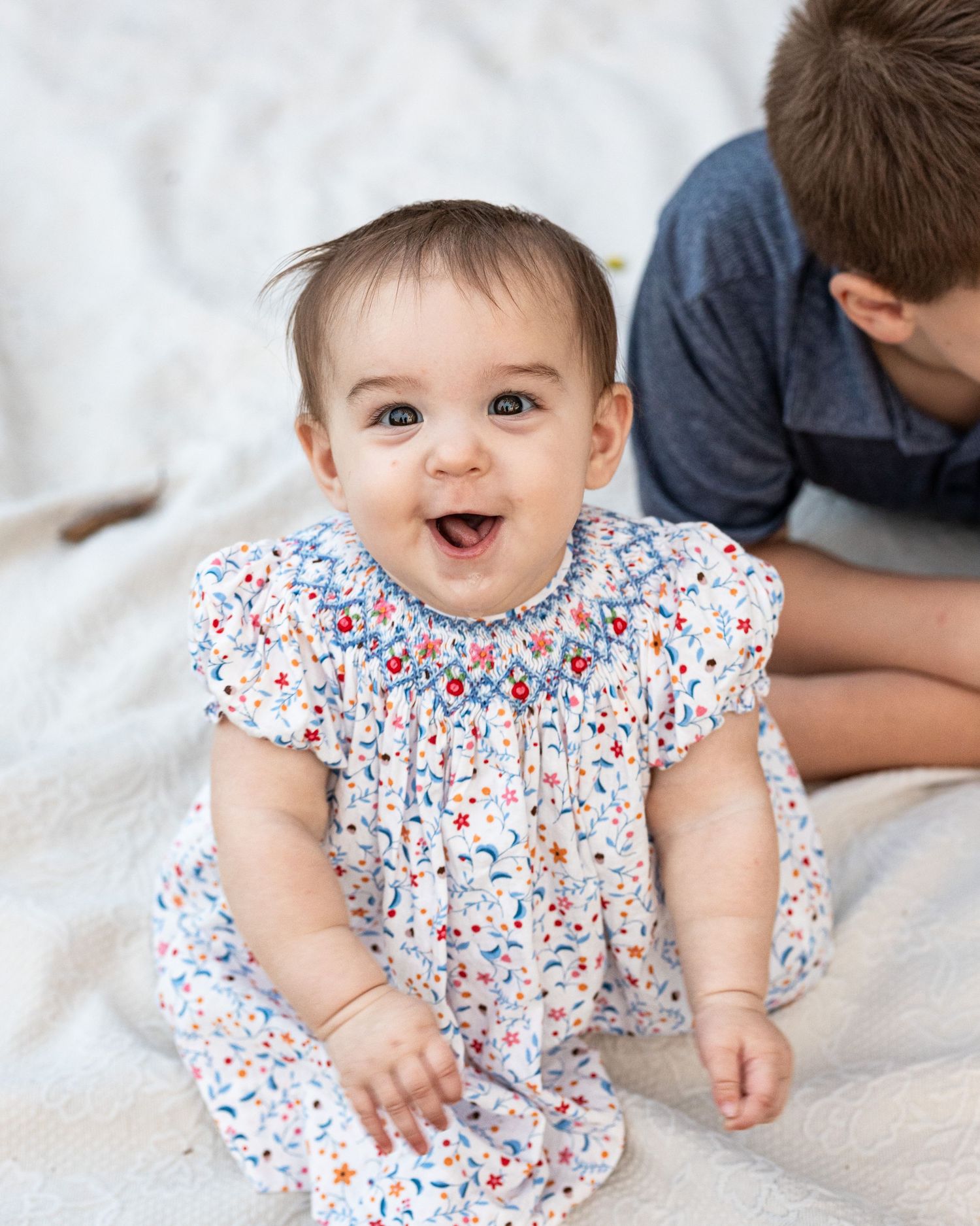 Baby in white and red floral dress lies on white bedding showing joyful expressions in progression.