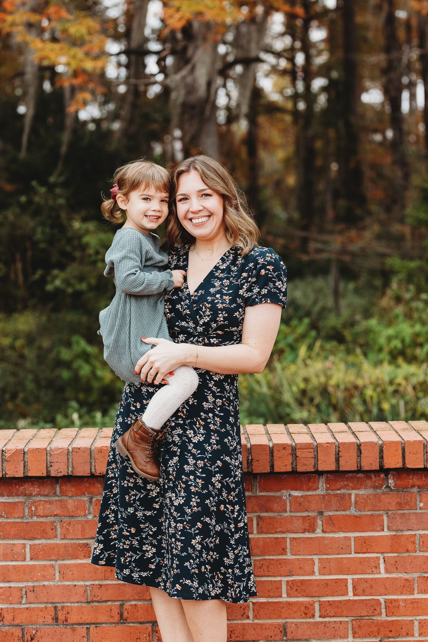 A fall portrait session featuring a floral dress against a red brick wall with autumn foliage in background.