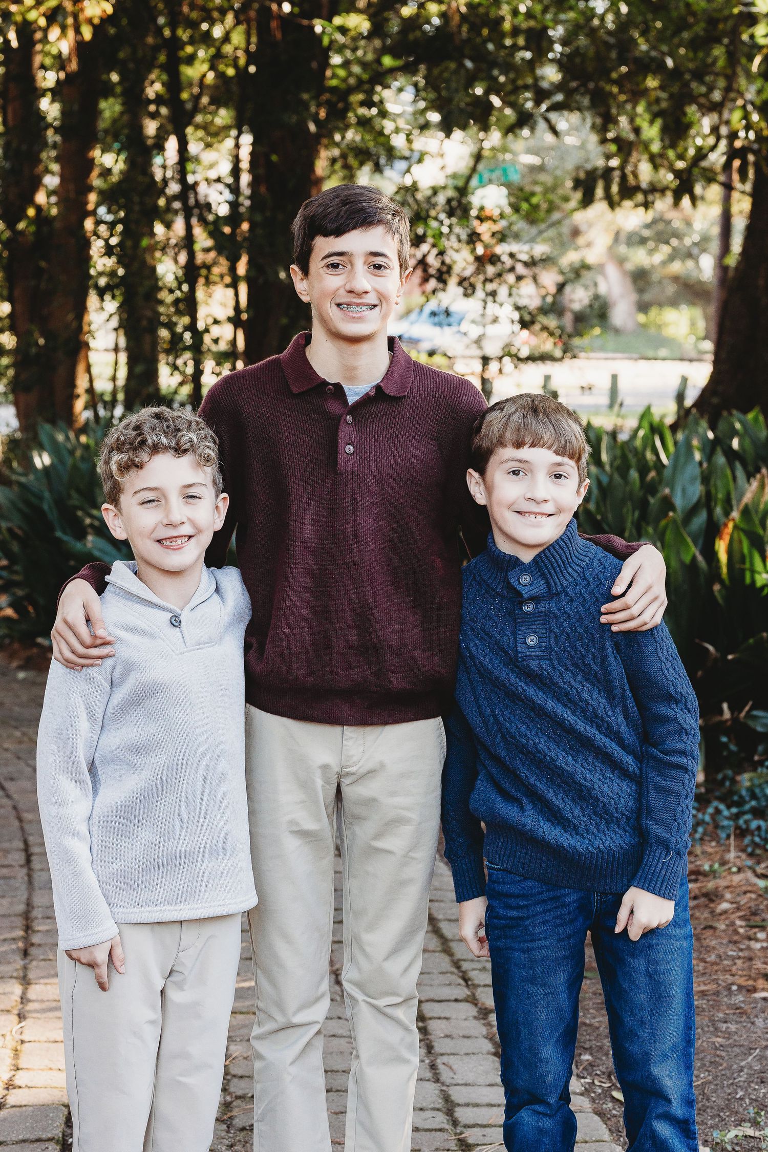 Three children in coordinating sweaters and khaki pants smile together in an outdoor setting.
