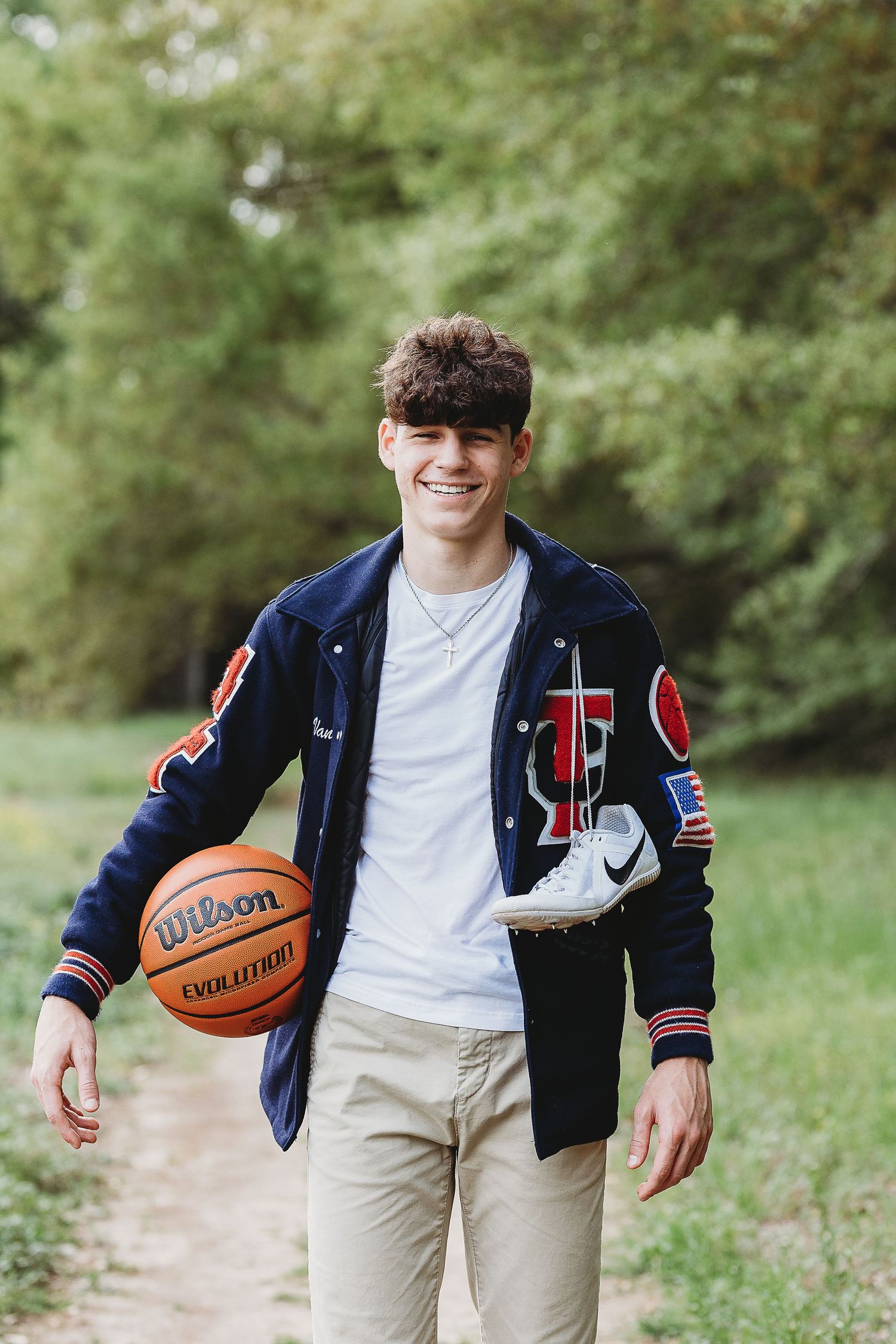 Person in varsity jacket holds basketball while posing on outdoor path surrounded by greenery.