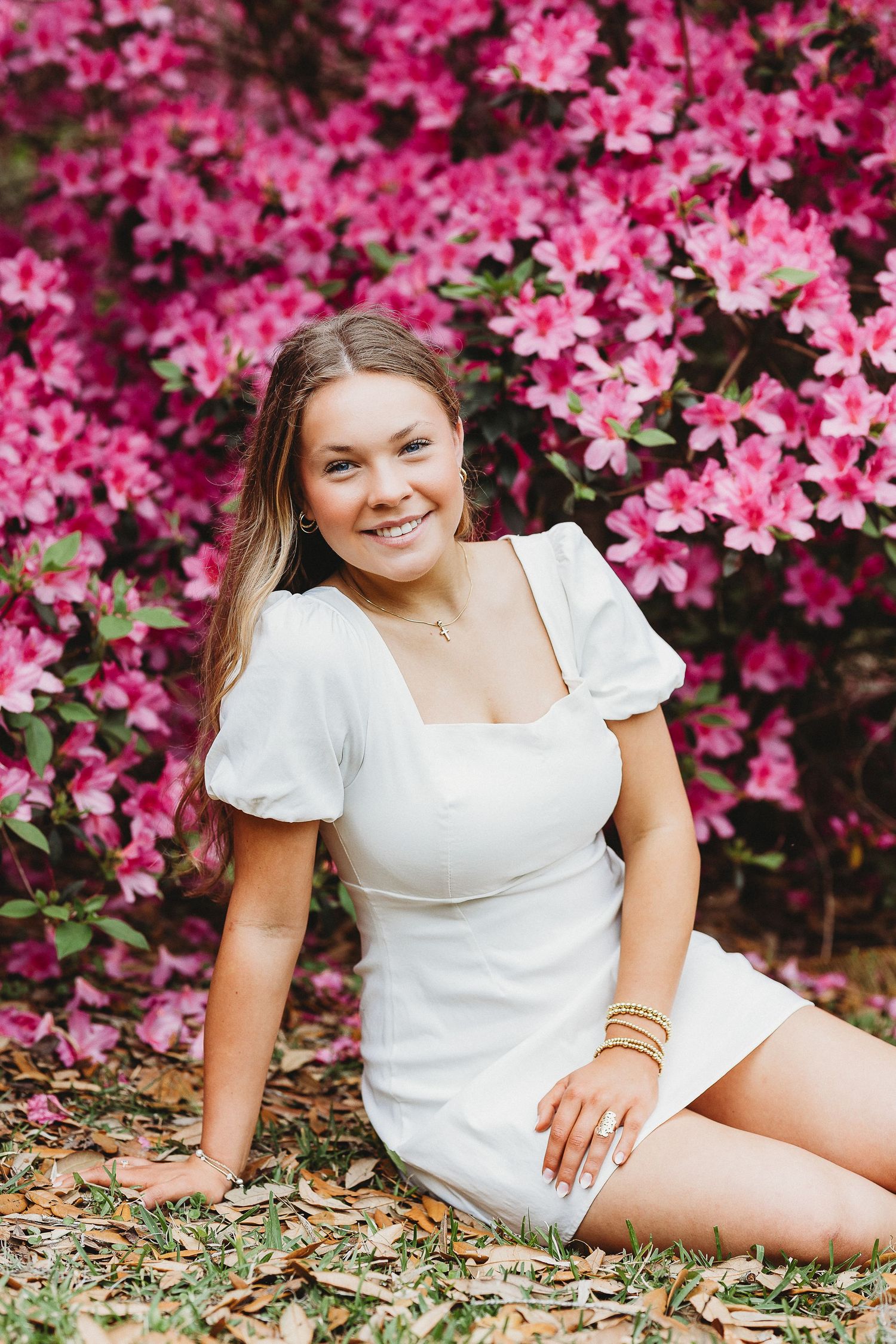 Person in white dress poses by bright pink flowering bushes during photo session.