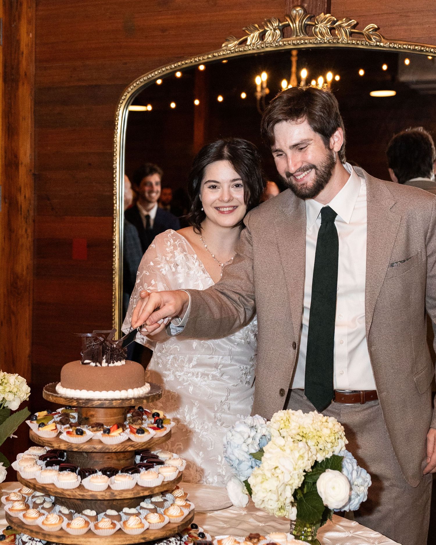 A wedding couple cuts into their rustic tiered chocolate wedding cake decorated with berries and flowers at their reception.