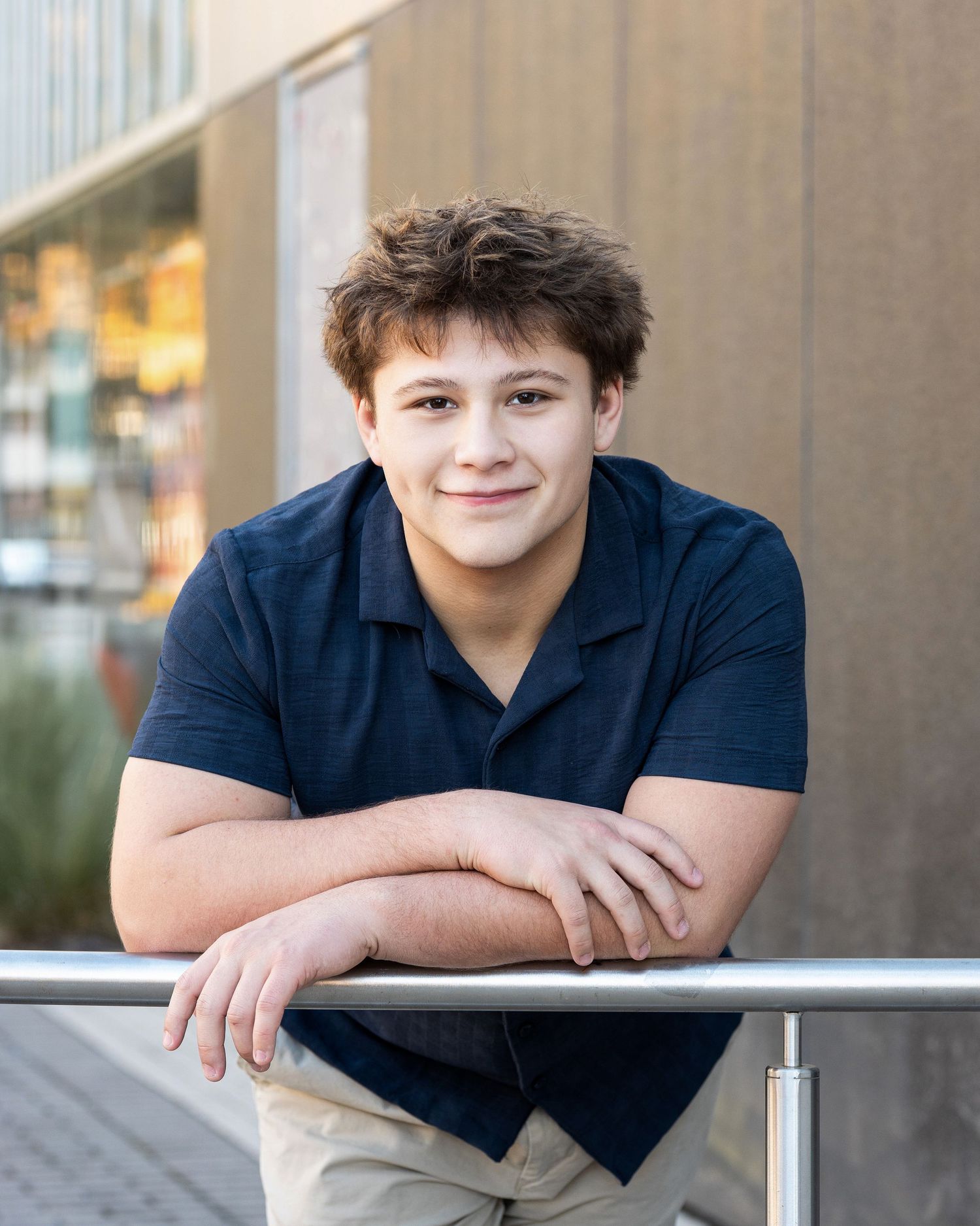 A young person in a navy polo shirt leans casually against a railing in an outdoor portrait setting.