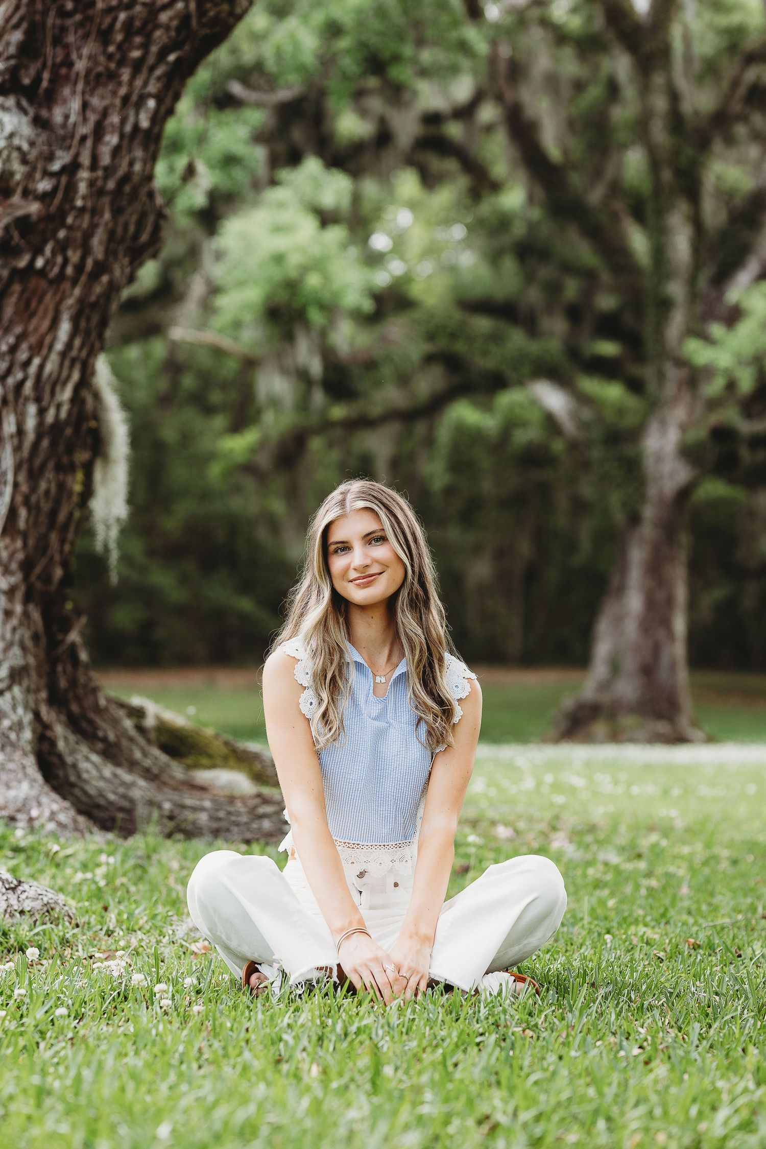 A person in a light blue top and white pants sits casually on lush green grass in a wooded park setting.
