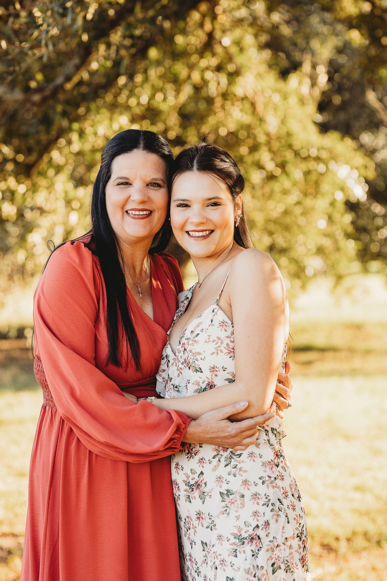 Two people share a warm embrace while wearing floral and coral dresses in golden sunlight.
