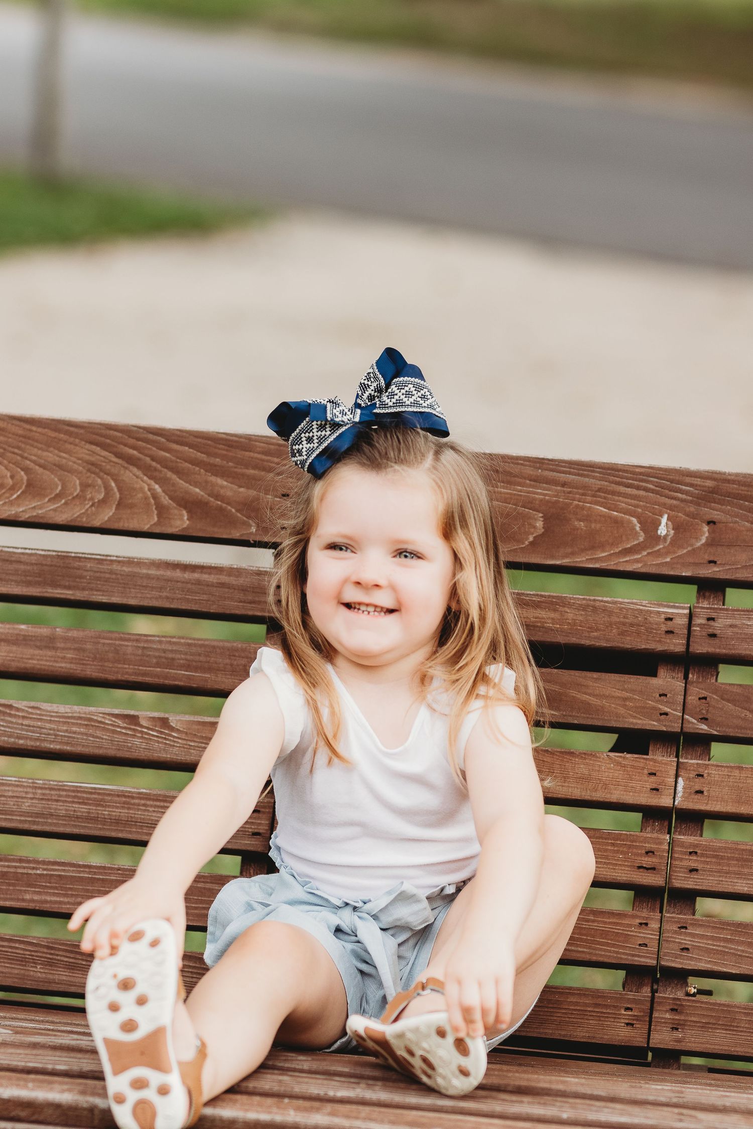 Young child in white tank top sitting on wooden bench with decorative blue bow.