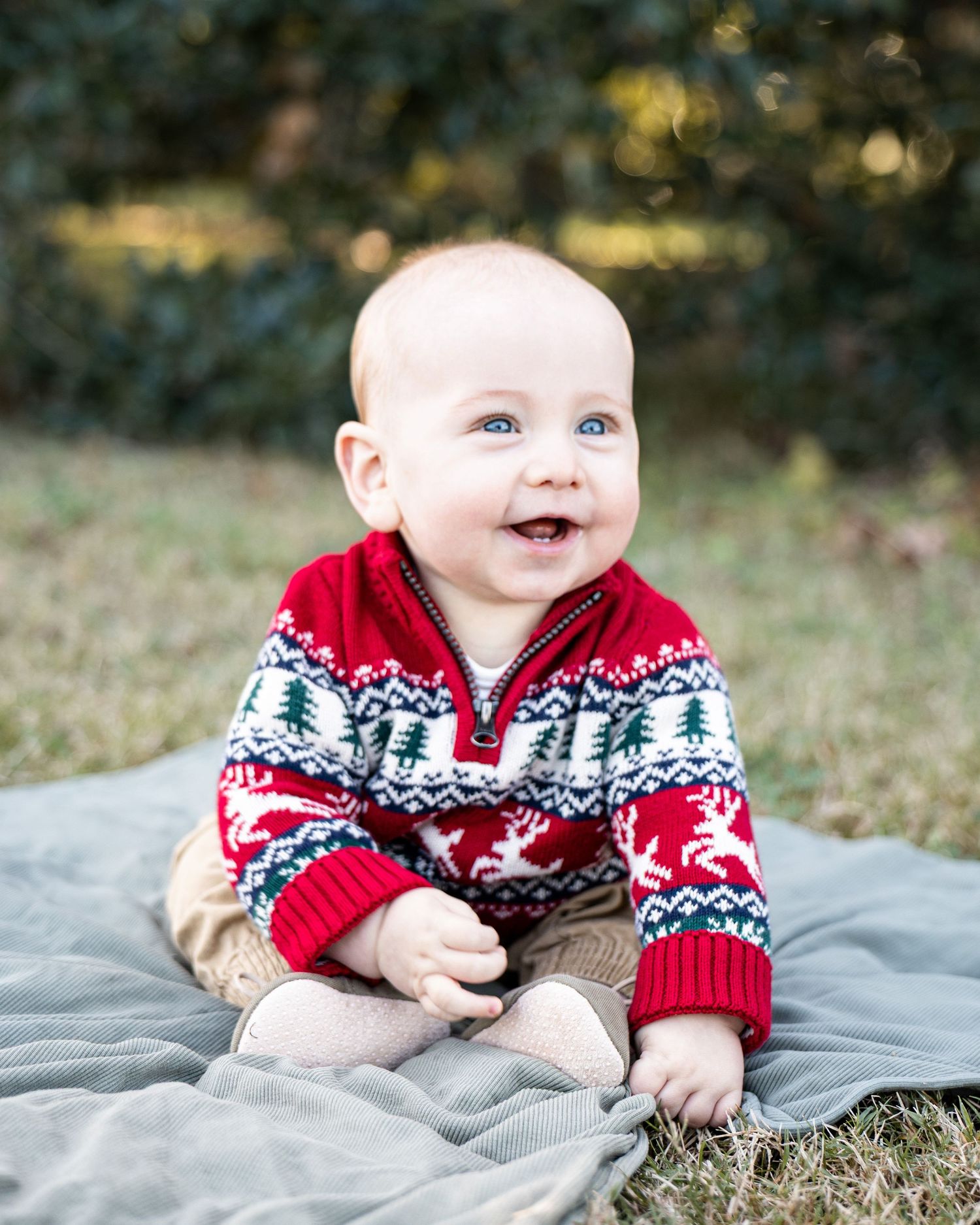 A baby wearing a festive red and white Nordic patterned sweater lies on a gray blanket outdoors.