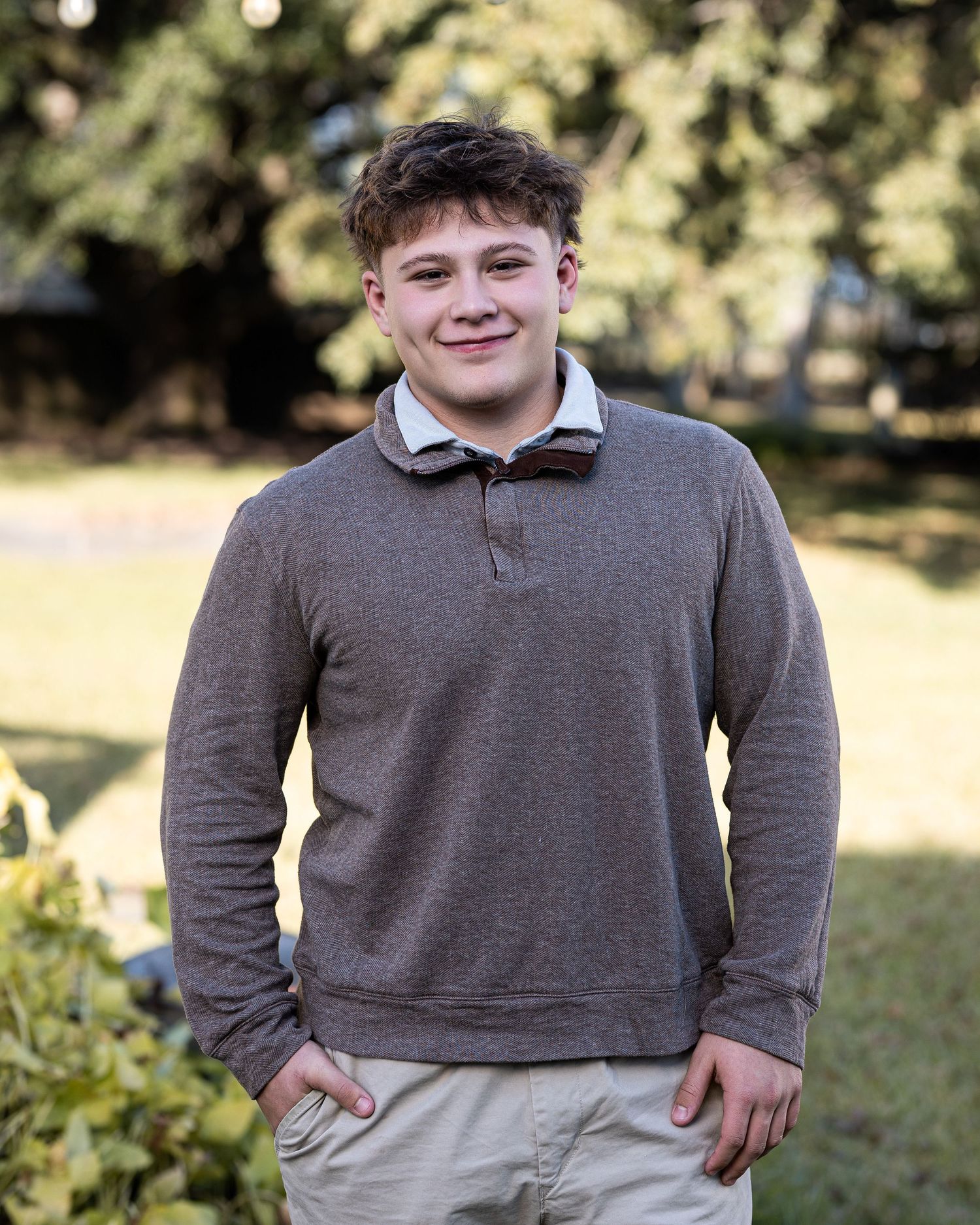 Young person wearing a gray collared shirt stands outdoors against a blurred natural background.