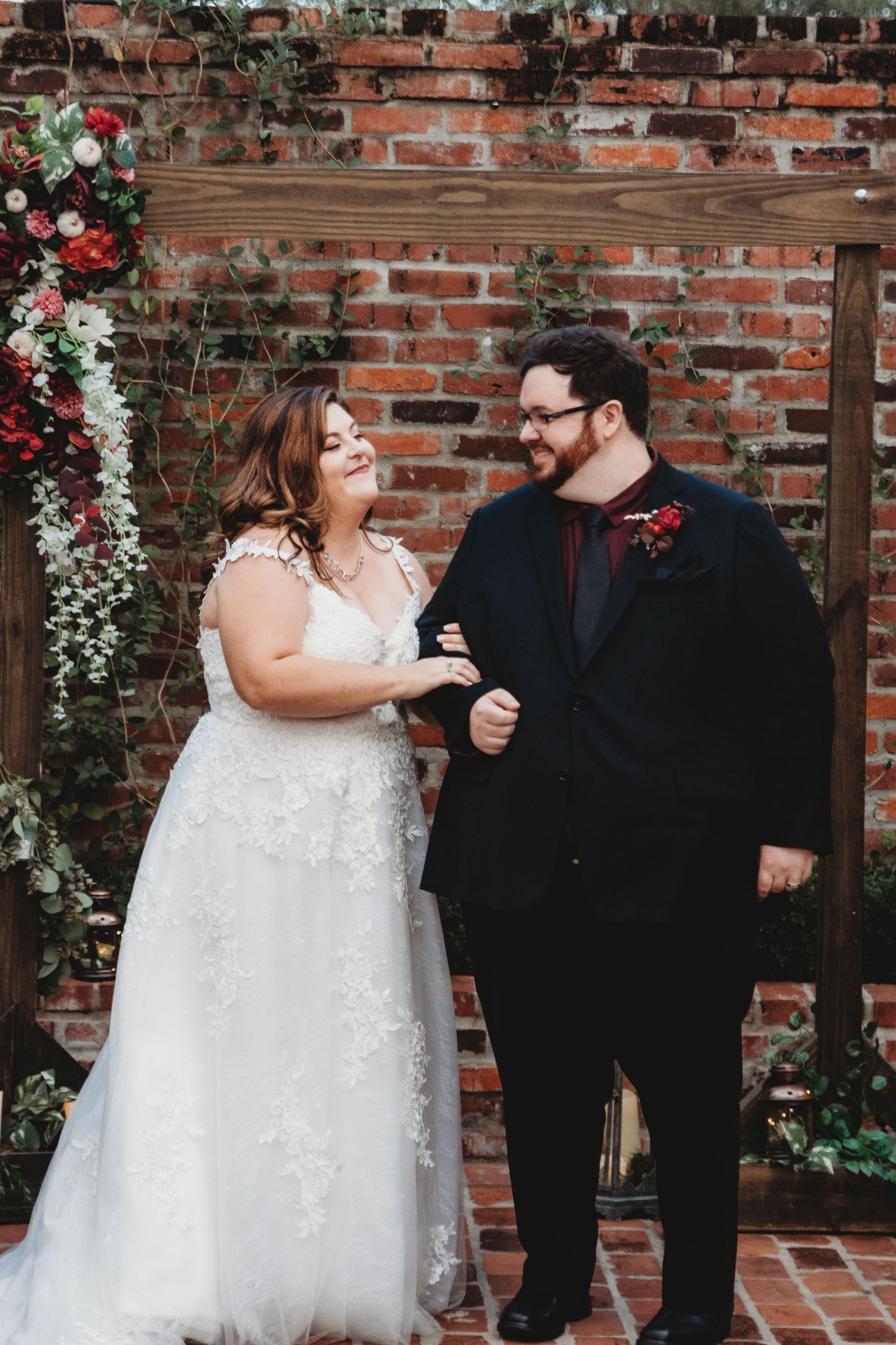Couple shares joyful moments against a rustic brick wall decorated with red and white flowers.