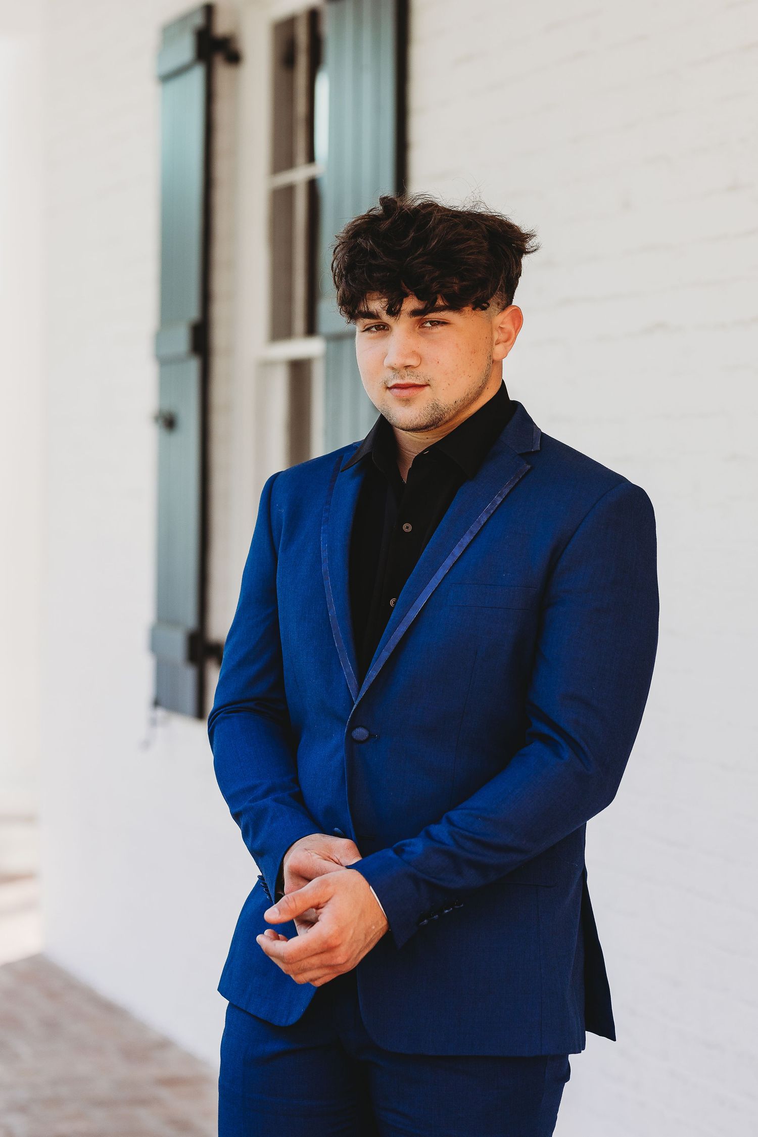 Formal portrait in navy blue suit and black shirt against white wall.