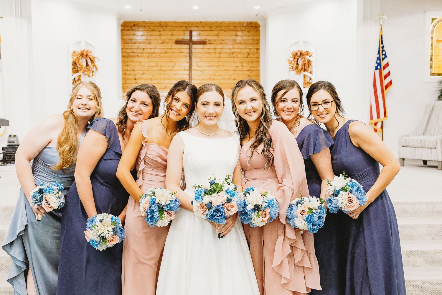 Bridesmaids in navy and pink dresses pose with bouquets featuring blue flowers in a church setting.