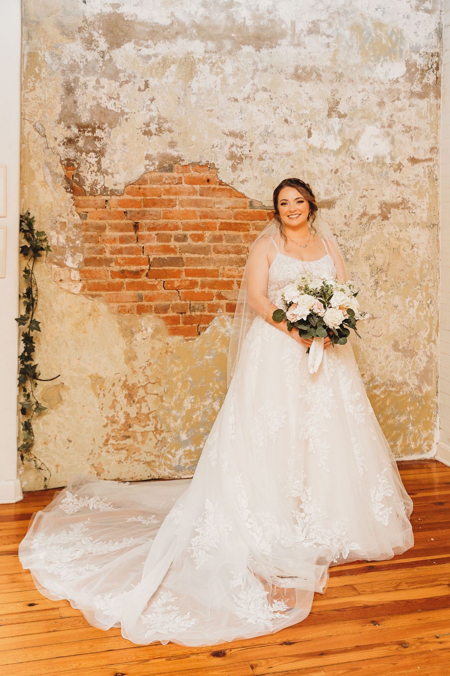 Bride in white wedding gown posing against rustic wall with exposed brick and wooden floor.