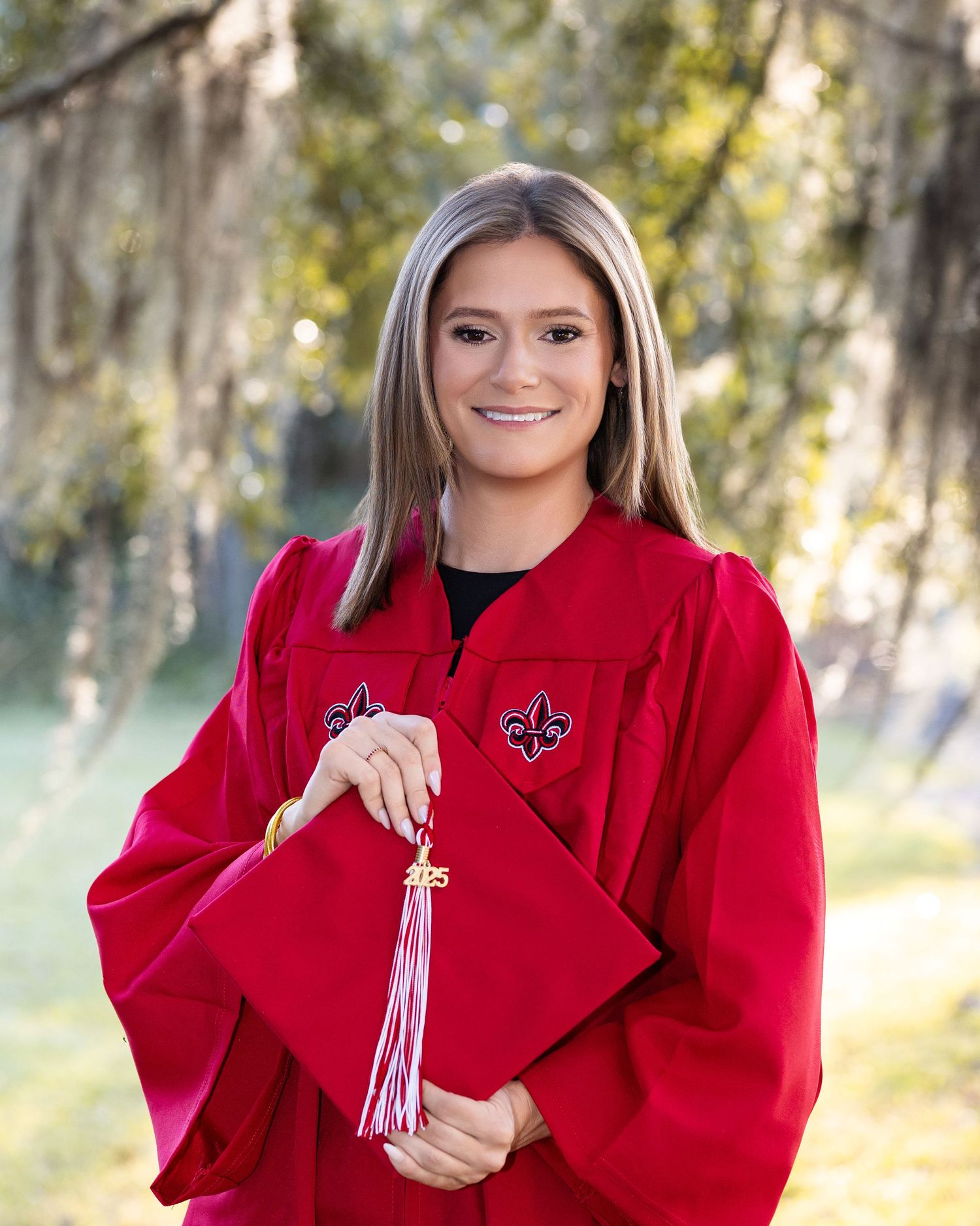 Graduate in red academic regalia holds white tassel while posing outdoors in natural sunlit setting.