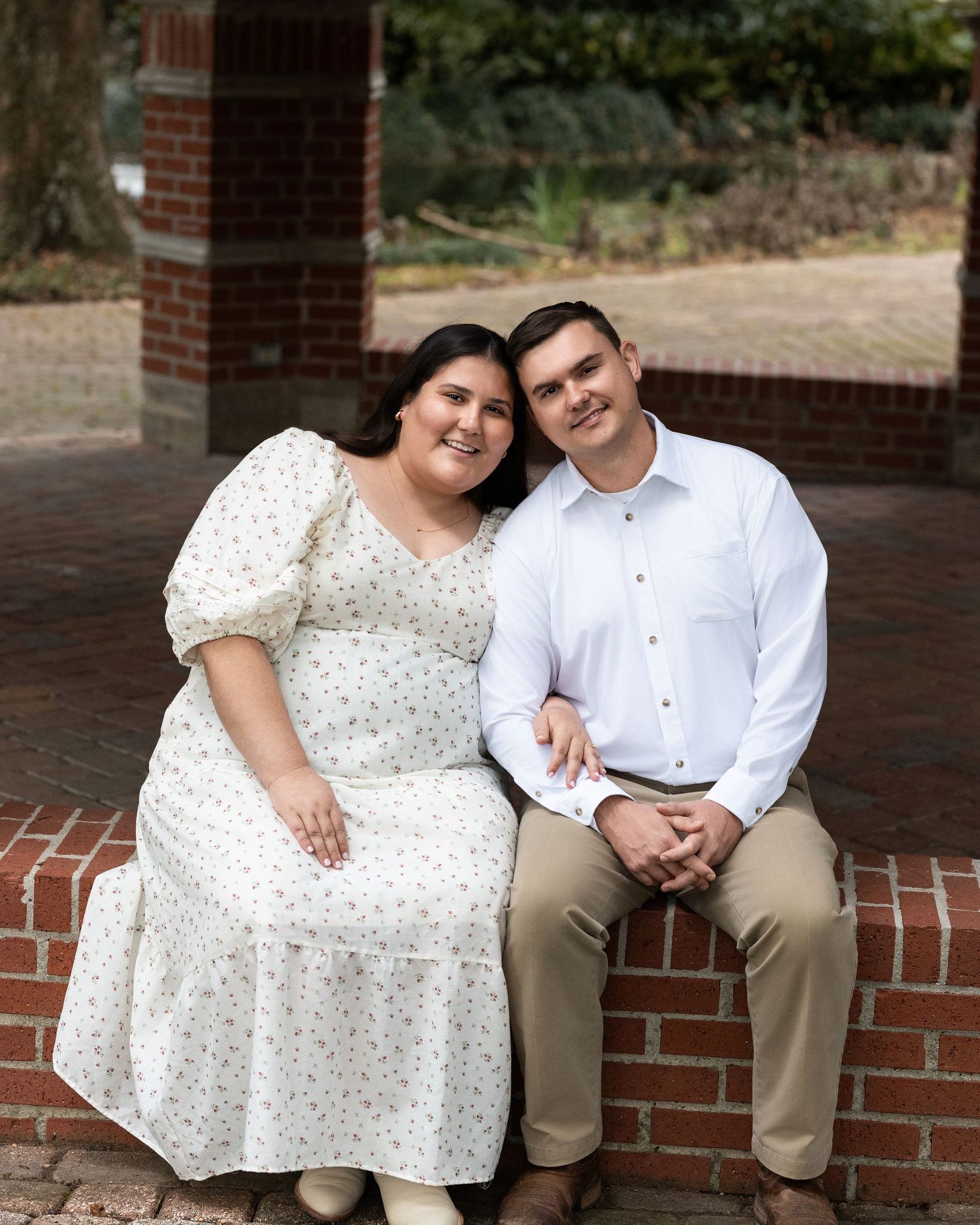 Couple sitting on brick ledge outdoors, wearing white polka dot dress and white button-down shirt.