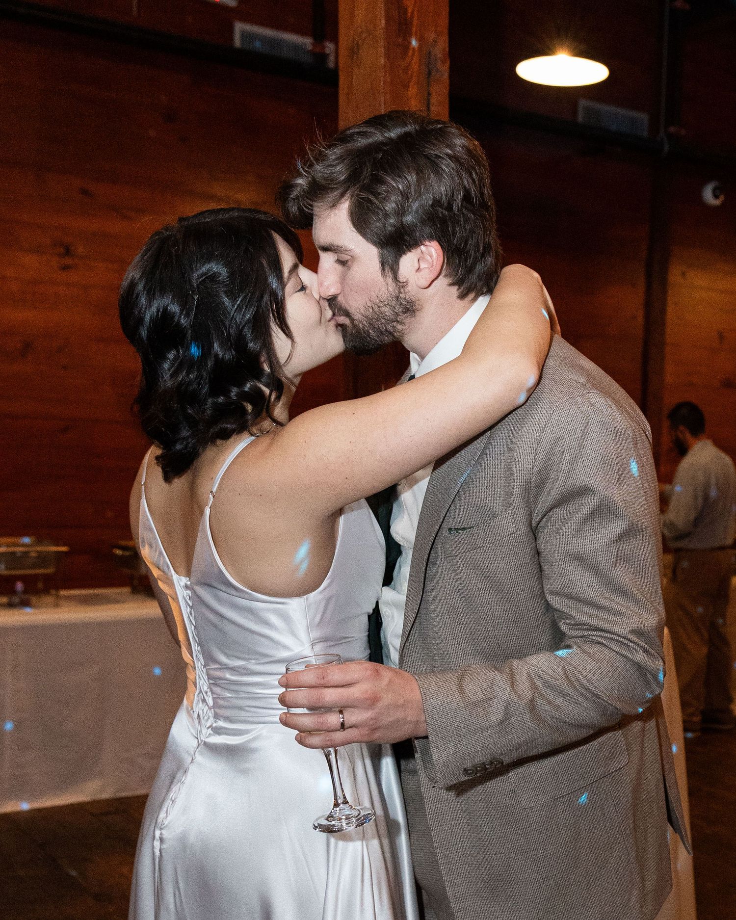 A couple in formal attire share a romantic kiss during their first dance at a rustic wooden barn wedding venue.