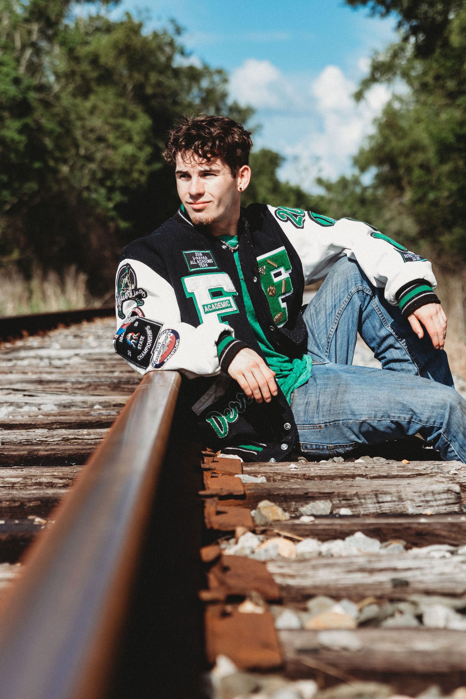A casual portrait of someone sitting on railroad tracks wearing a green varsity jacket and jeans.