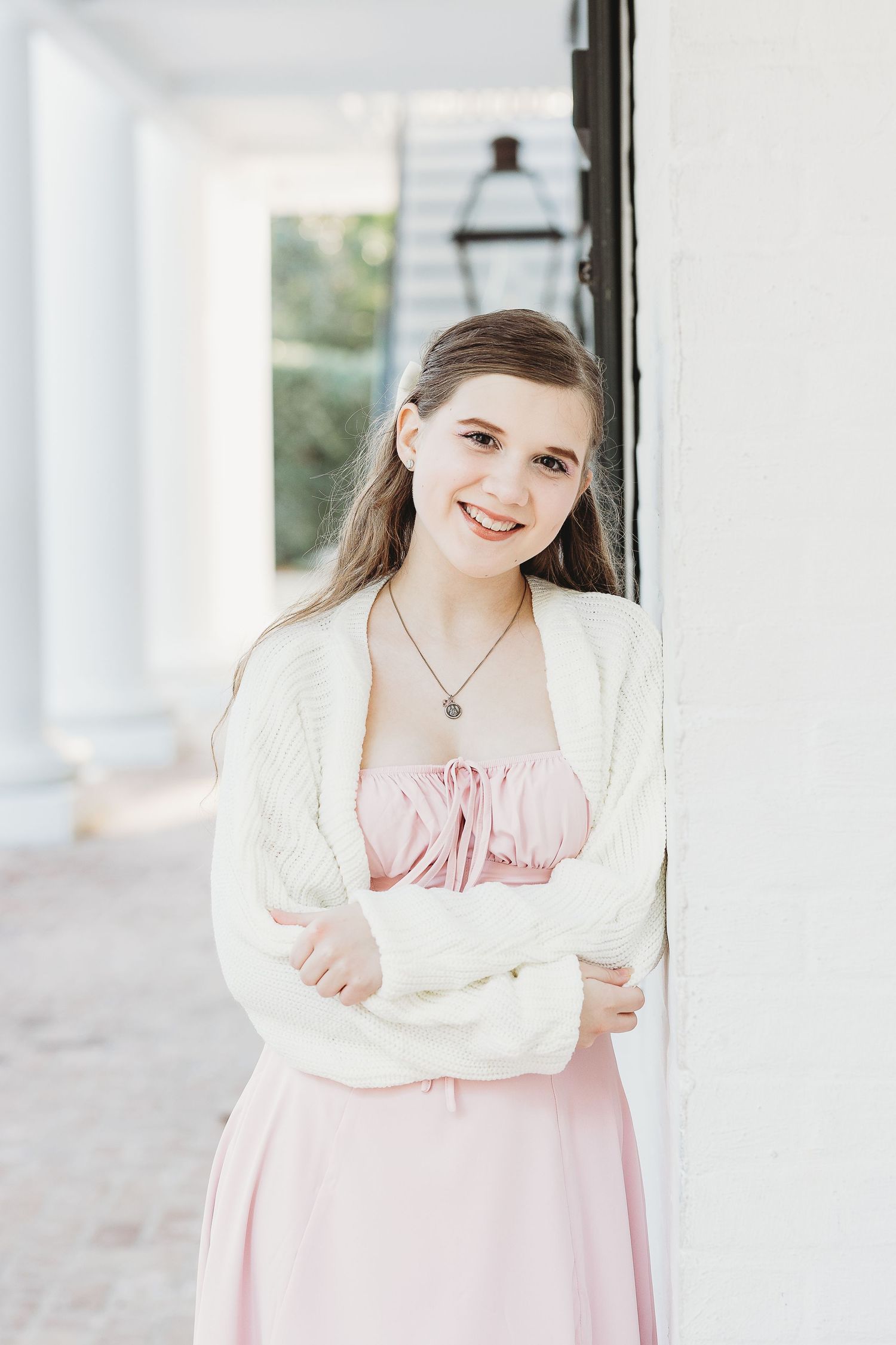 Person in pink dress and white cardigan poses elegantly against white column backdrop.