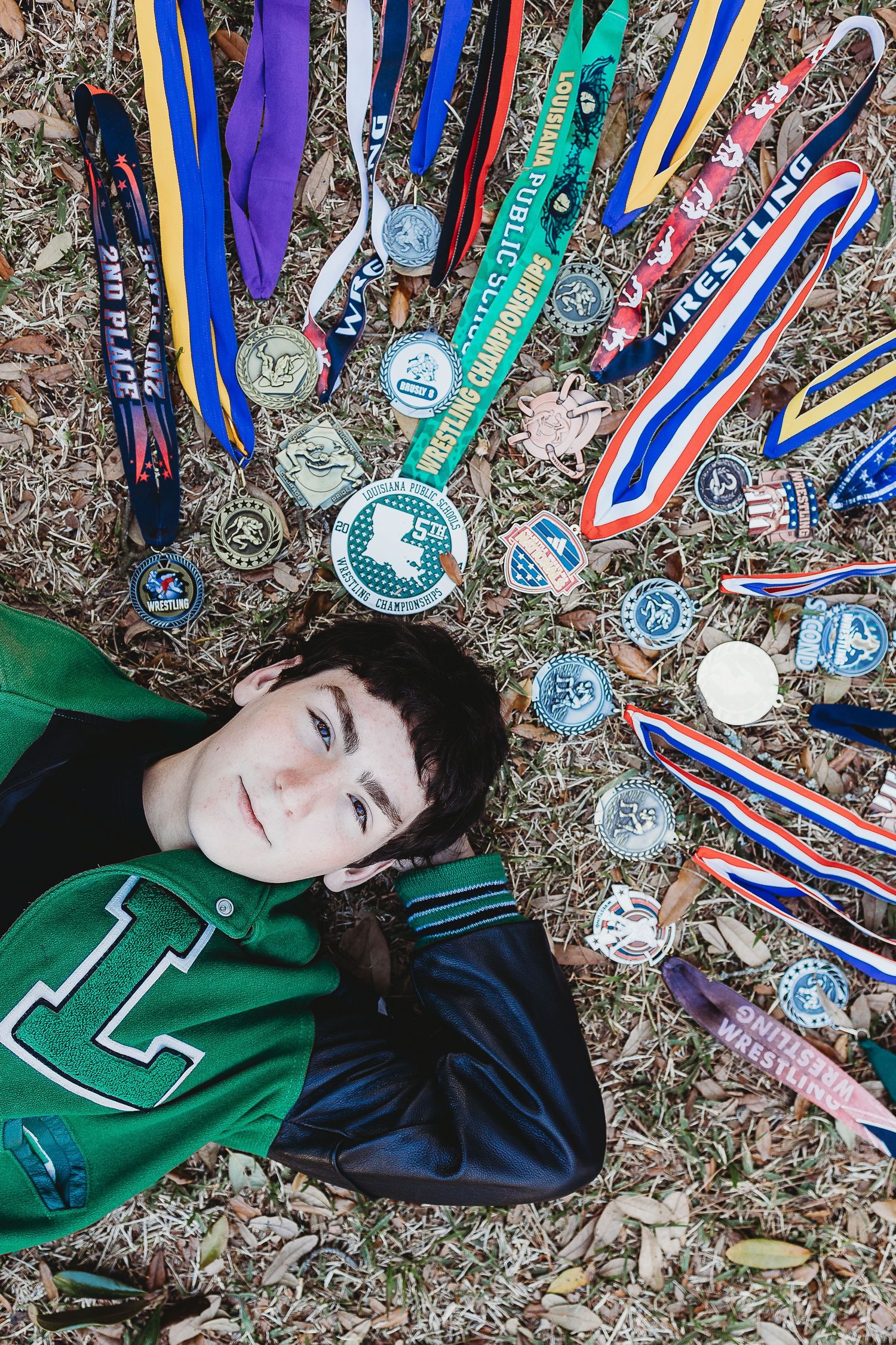 Athletic medals and ribbons displayed around a student in a green letterman jacket lying on fallen leaves.