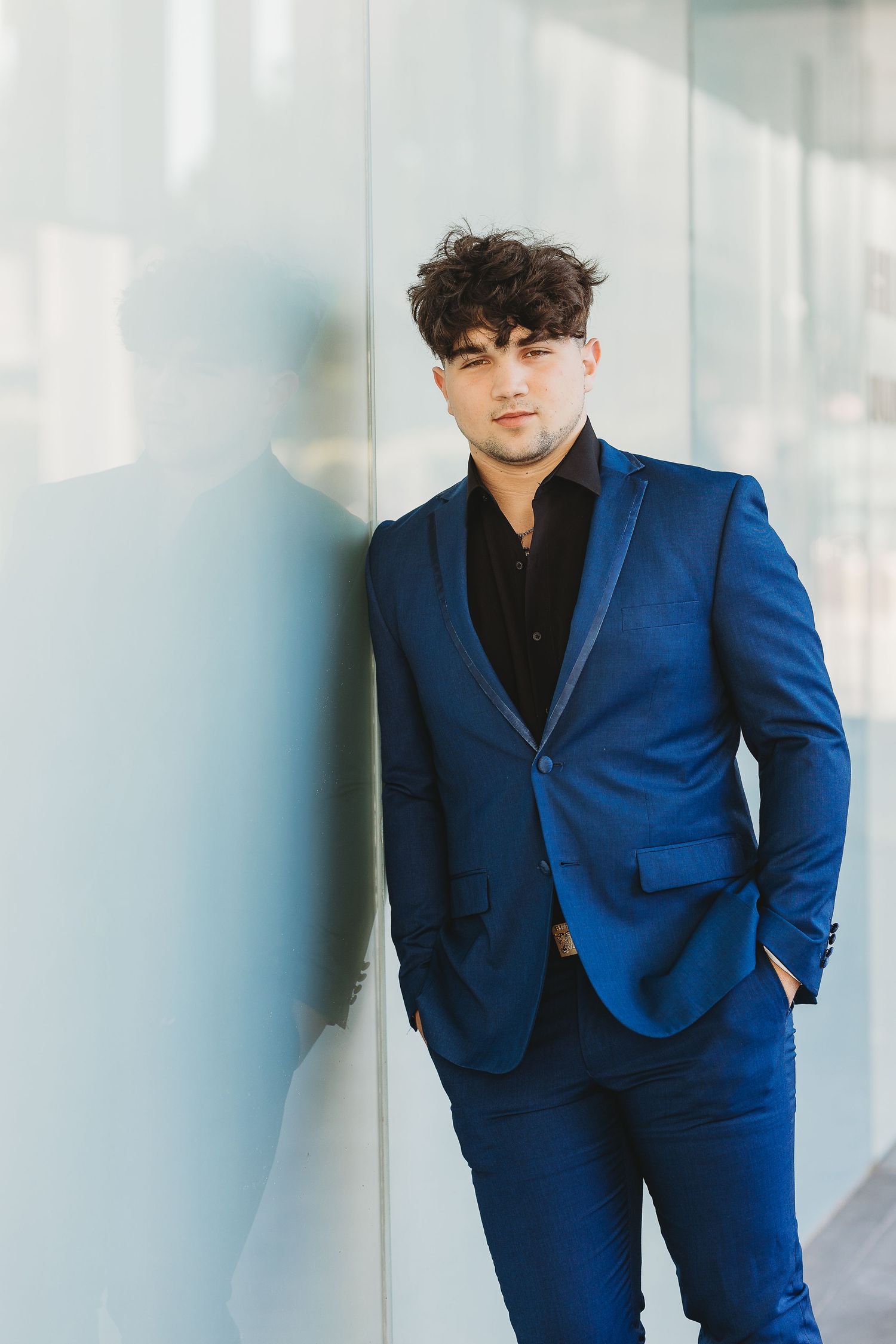 Professional portrait of someone in a fitted navy blue suit and black shirt against a light wall.