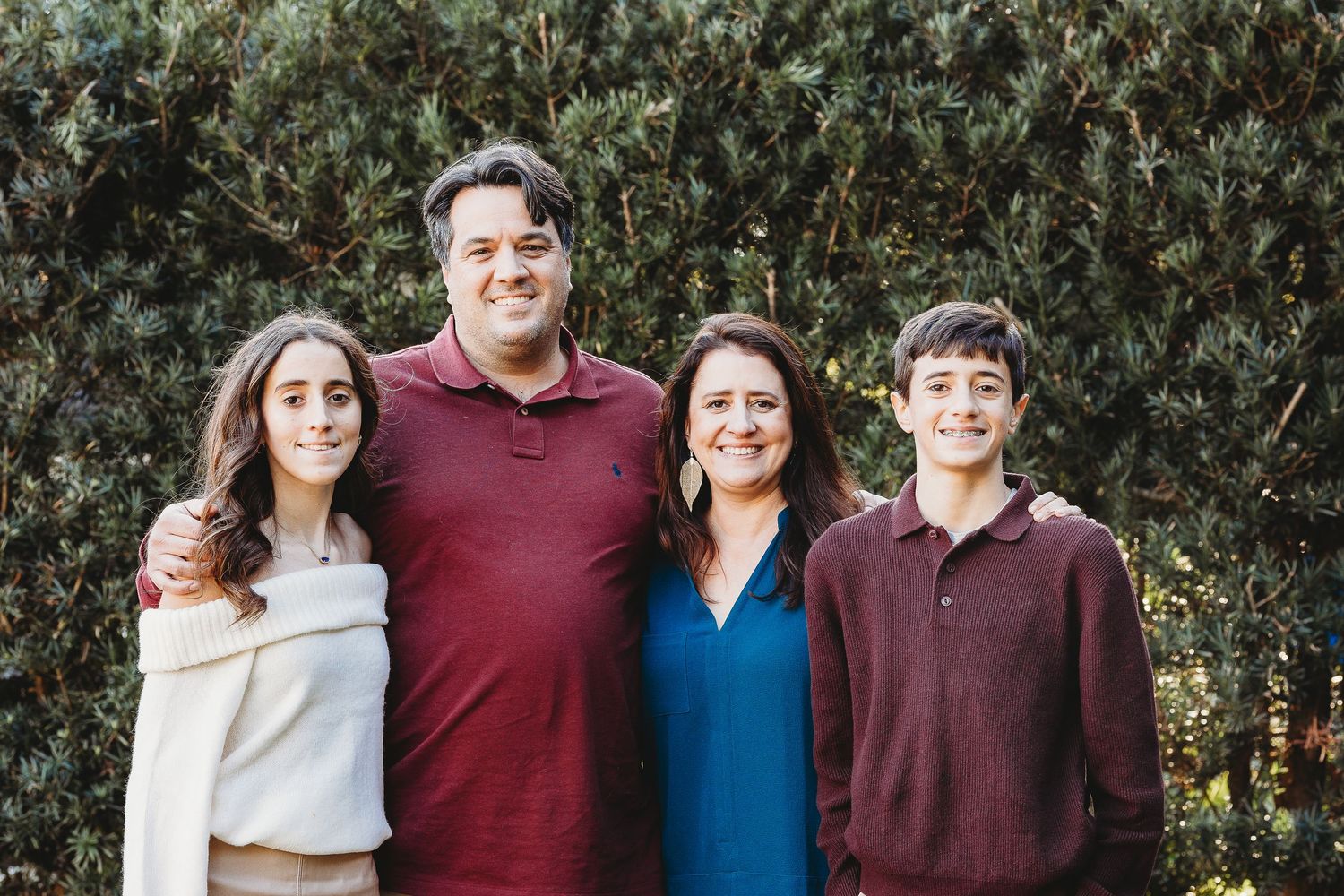 Family portrait showing multiple people in coordinating burgundy and blue outfits against natural backdrop.