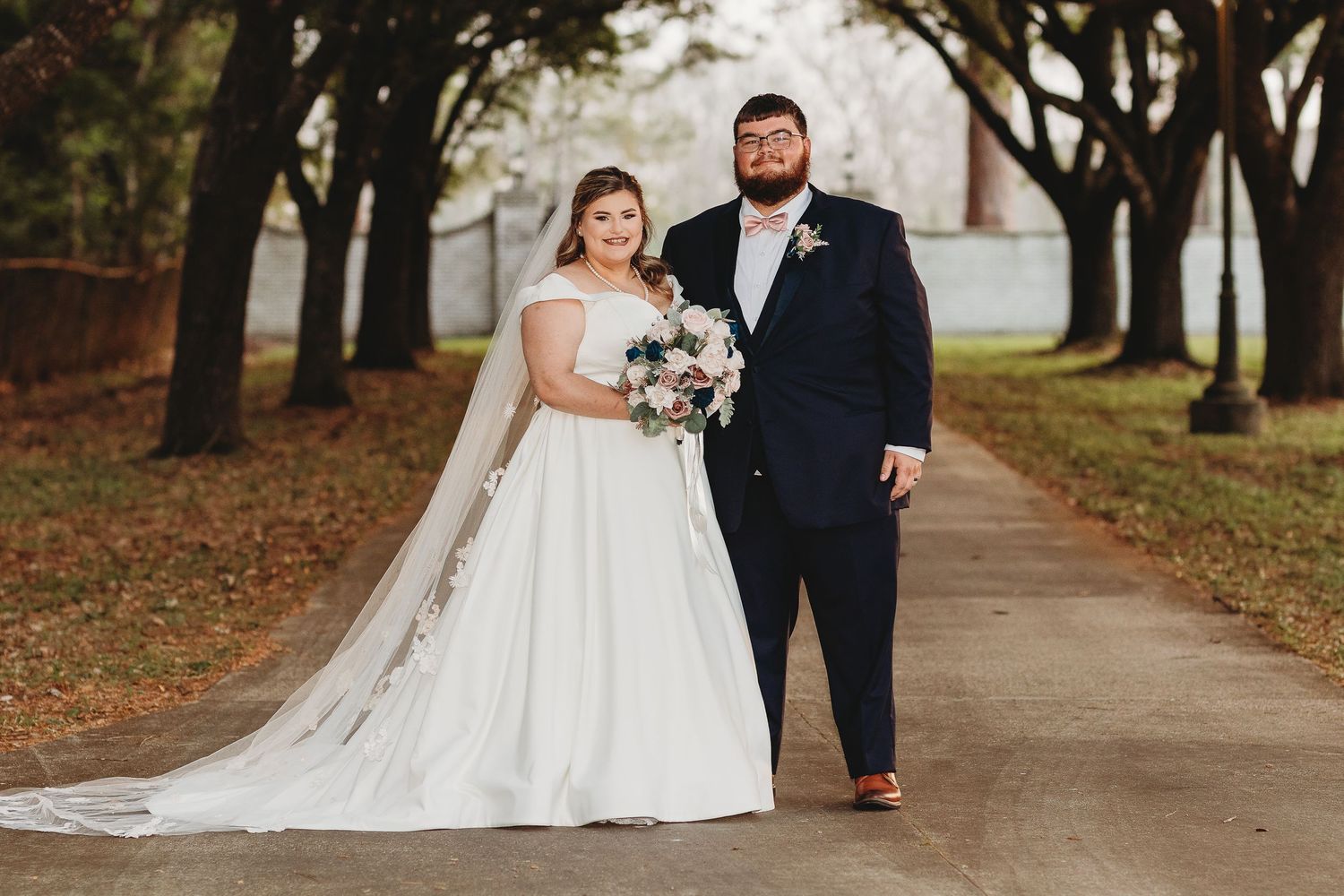 Wedding couple poses along tree-lined pathway during autumn outdoor photo session.