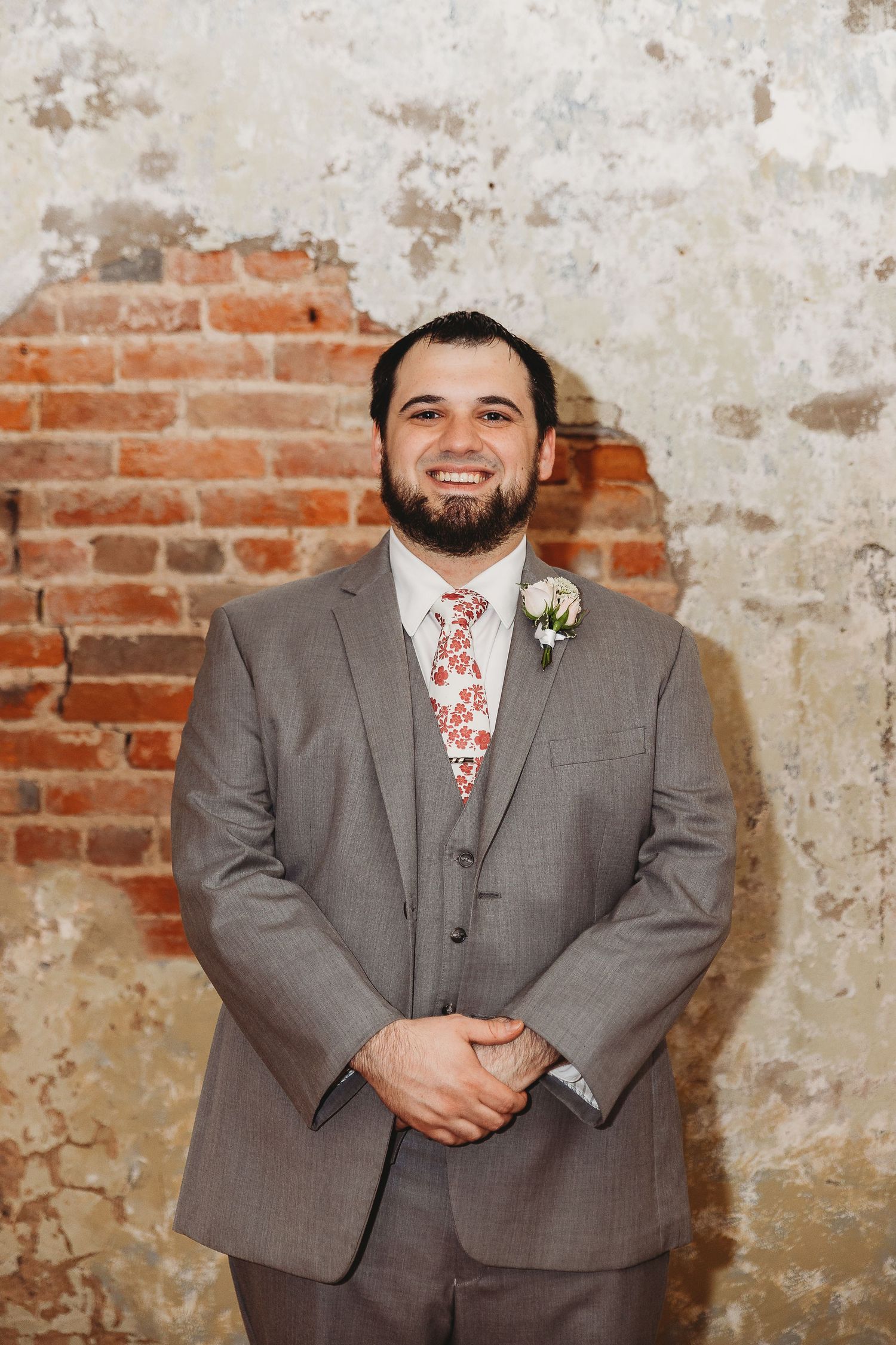 Groom in a grey suit with floral tie standing against a rustic brick and plaster wall.