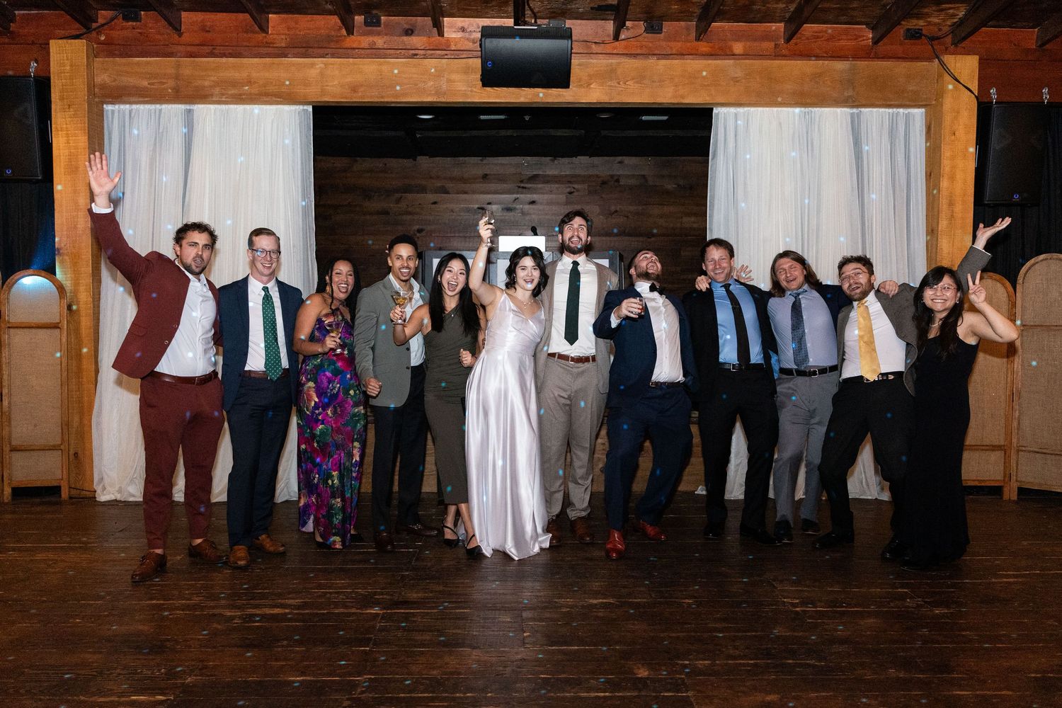 A wedding party poses together with raised arms in celebration inside a rustic wooden barn venue.