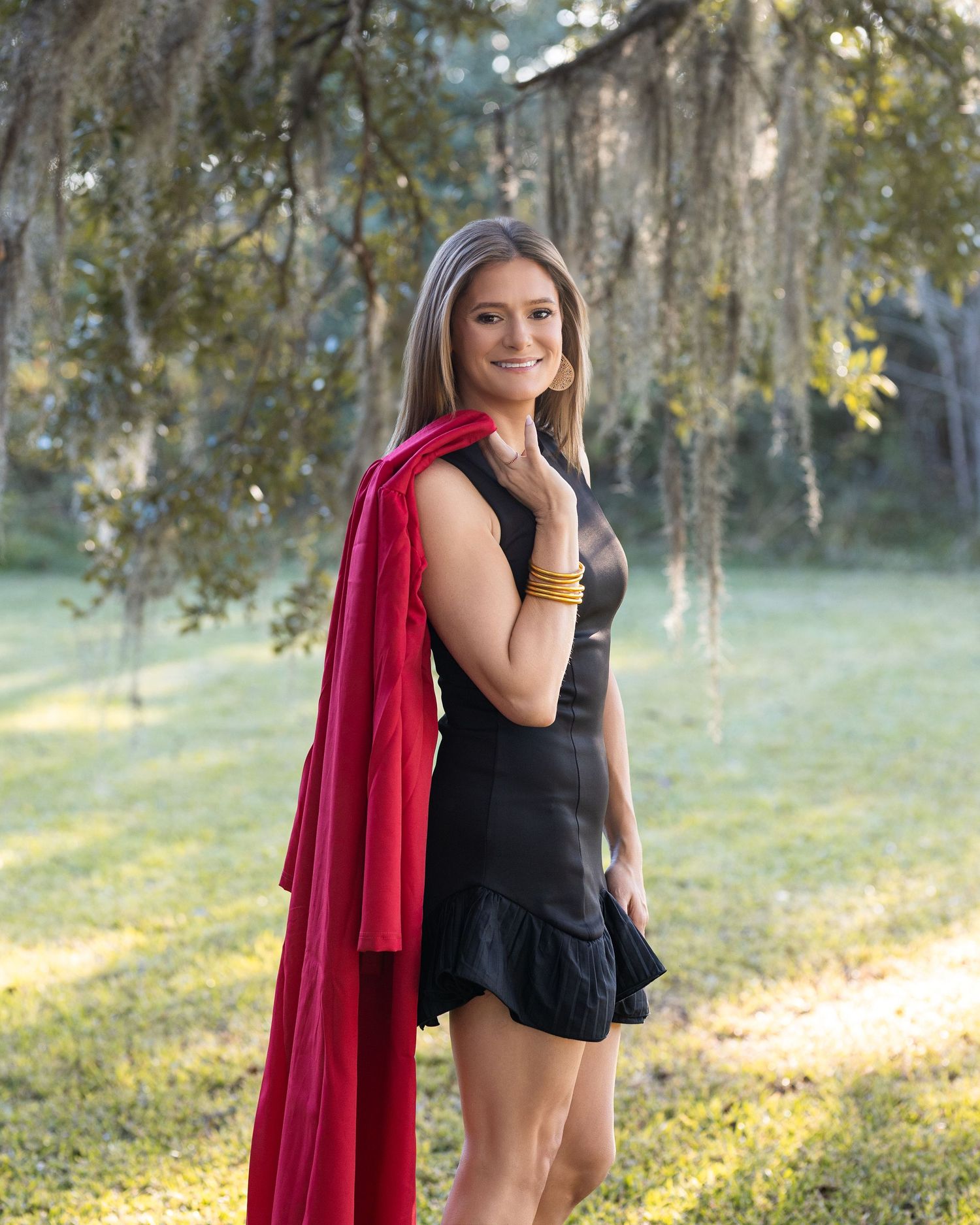 A person in a black romper and flowing red cape poses gracefully in a sunlit forest setting.